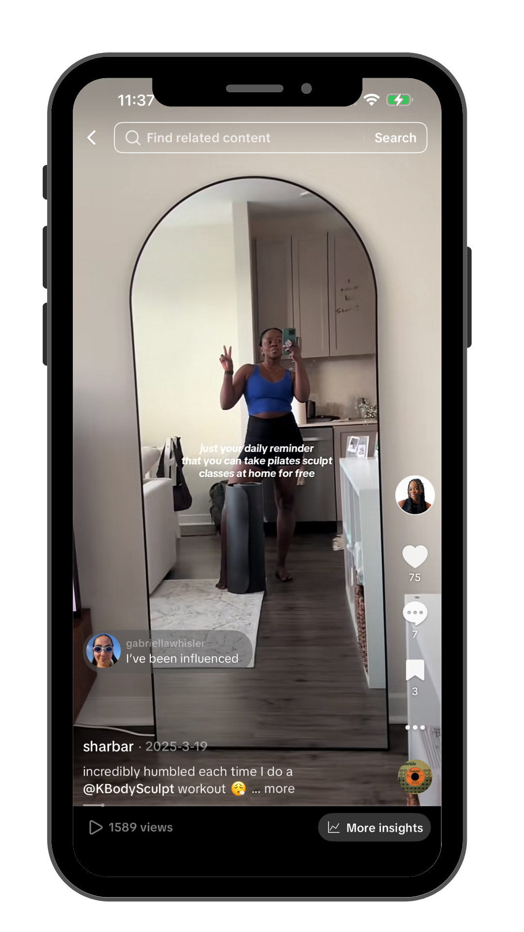 A woman is taking a mirror selfie in a living space, making a peace sign with her right hand, wearing a blue sports bra and black shorts. The mirror is arched, and the background shows a kitchen area with cabinets and a countertop.