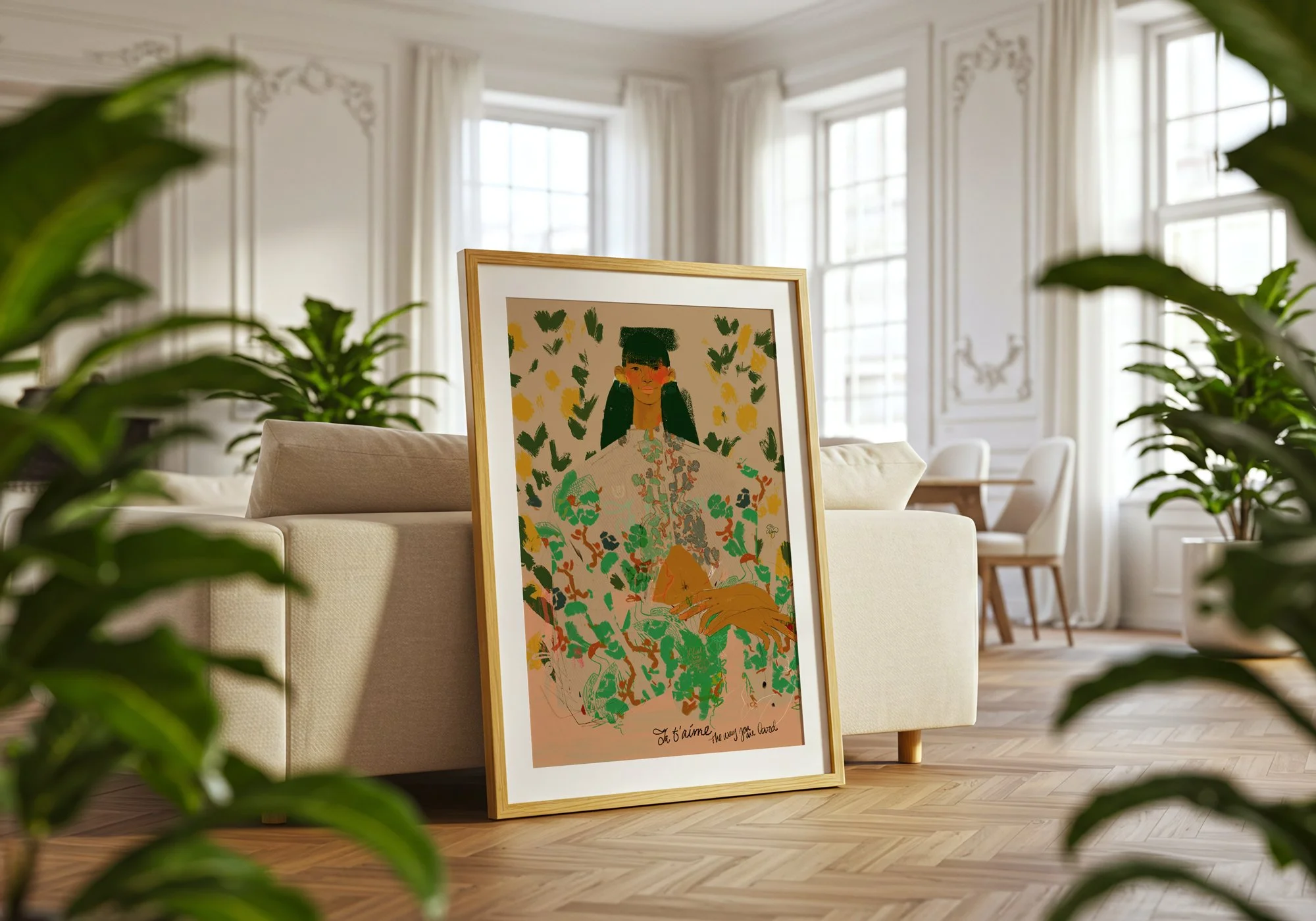 A colorful framed art print leaning against a beige sofa in a bright living room with large windows, white walls, and potted plants.