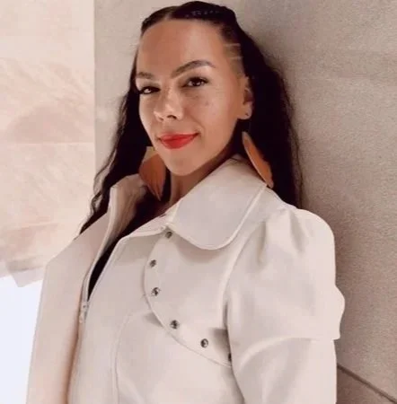 A woman with long dark hair, wearing a cream-colored jacket and orange earrings, standing against a beige wall.