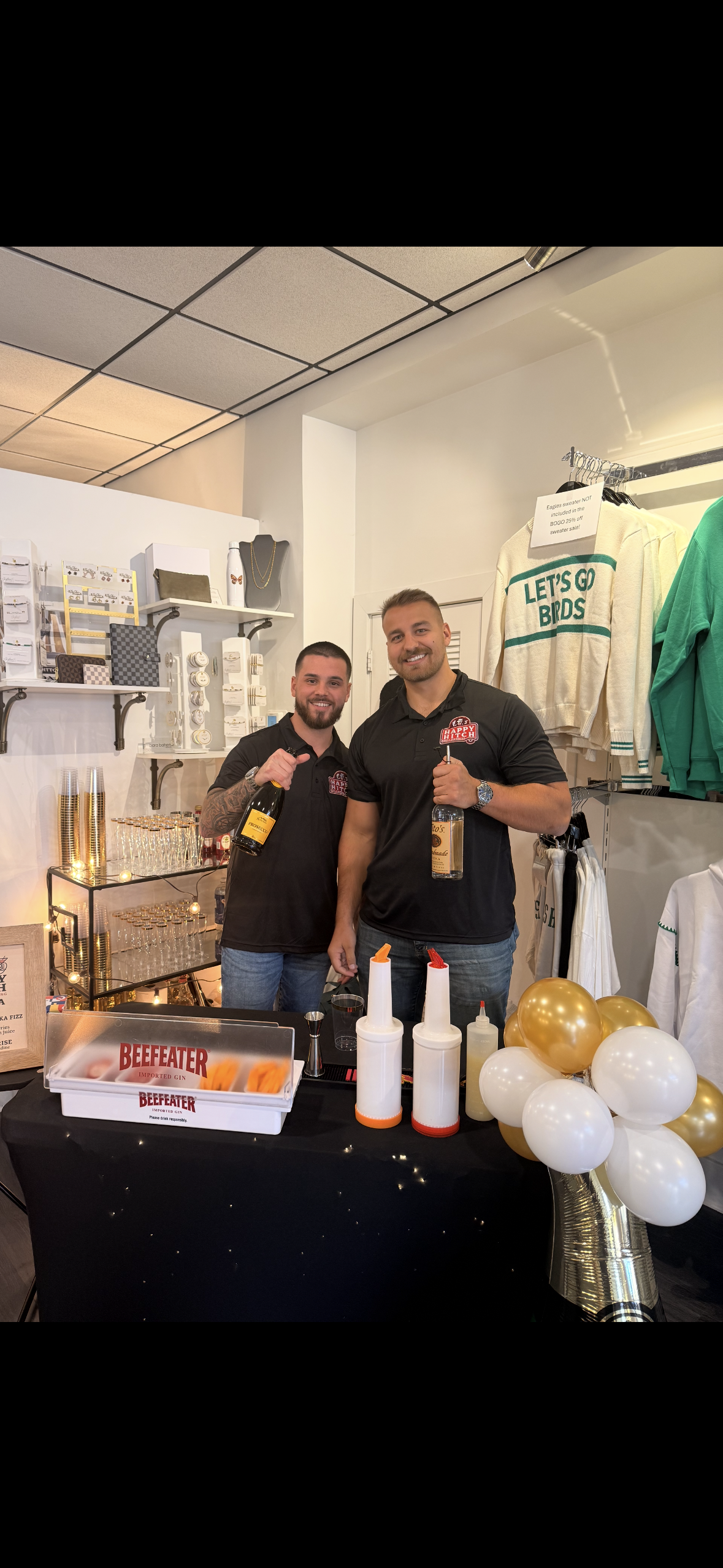 Two men standing behind a black table with balloons, condiments, and a Beefeater gin box, smiling and holding bottles inside a retail store or event space.