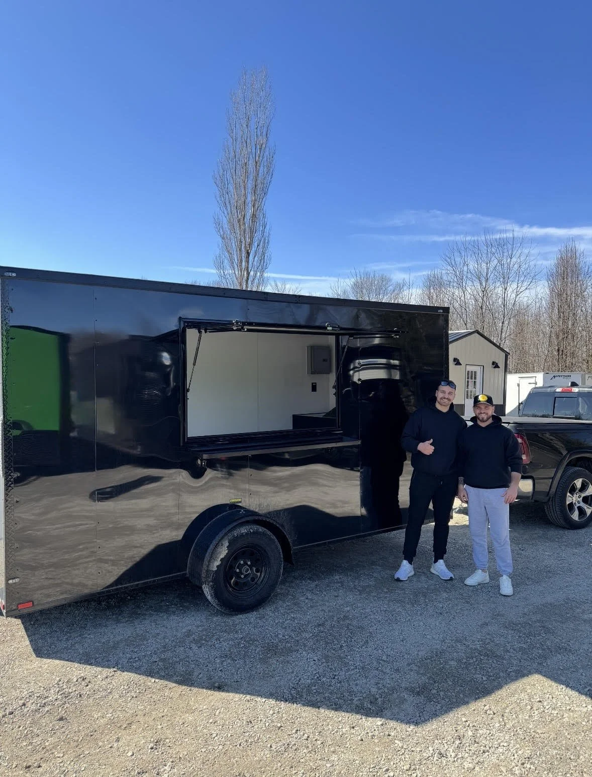 Two men standing outside next to a black food truck and a black pickup truck on a gravel lot under a clear blue sky.