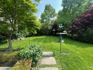 A lush green backyard with a well-maintained lawn, surrounded by trees and shrubs, with a stone pathway and bird feeders.