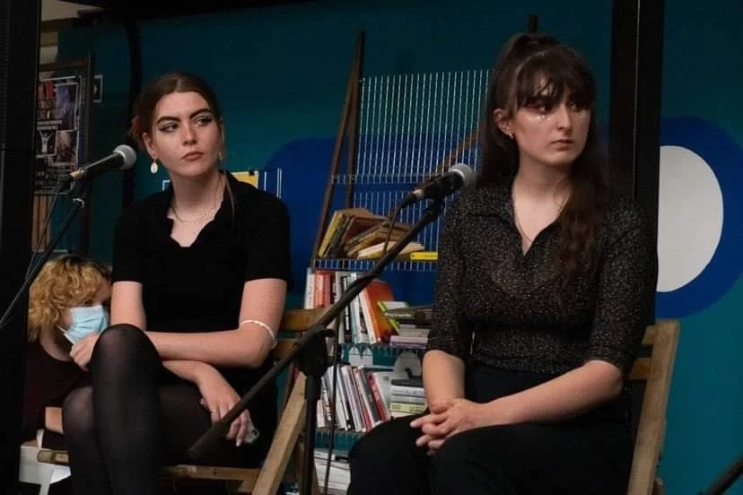 Two young women sitting in front of microphones at a panel or discussion event, with books and a blue wall in the background. One woman has dark hair and is wearing a black top, the other has long dark hair, wearing a black patterned top. A young girl in a face mask is partially visible on the left.