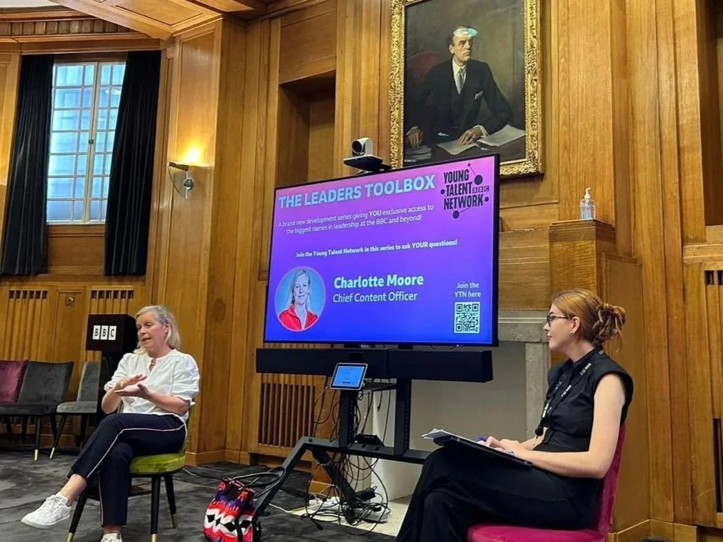 A woman and a girl sitting and talking in a cozy, wood-paneled room with a large window. There is a large digital screen displaying a presentation titled 'The Leaders Toolbox' with a photo of Charlotte Moore, Chief Content Officer, and QR code. There are chairs, a small table, and a bottle of hand sanitizer on a shelf.