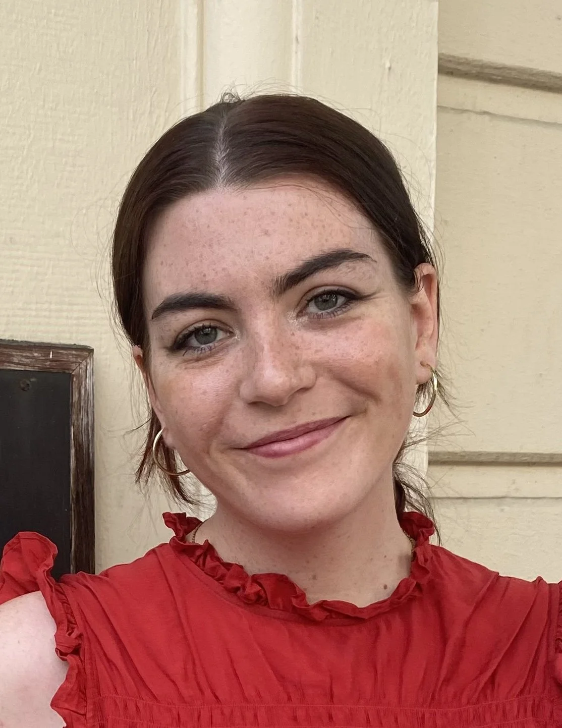 A young woman with dark brown hair, green eyes, and fair skin with freckles, smiling, wearing a red ruffled top, standing in front of a cream-colored wall with some dark accents.