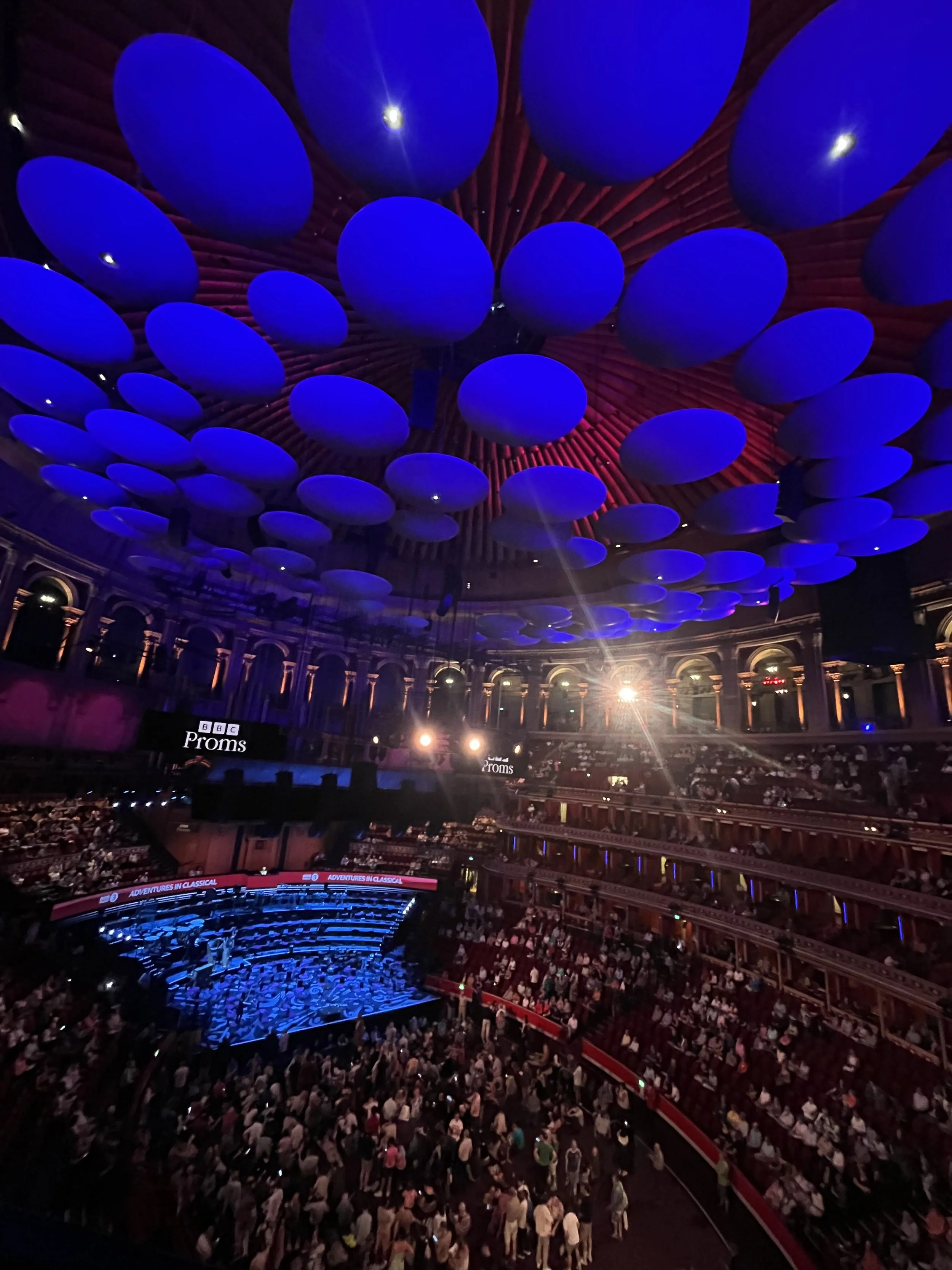 Concert hall with an ornate ceiling decorated with large blue circular acoustic panels, a stage with the BBC Proms logo, and a large audience seated in multiple tiers.
