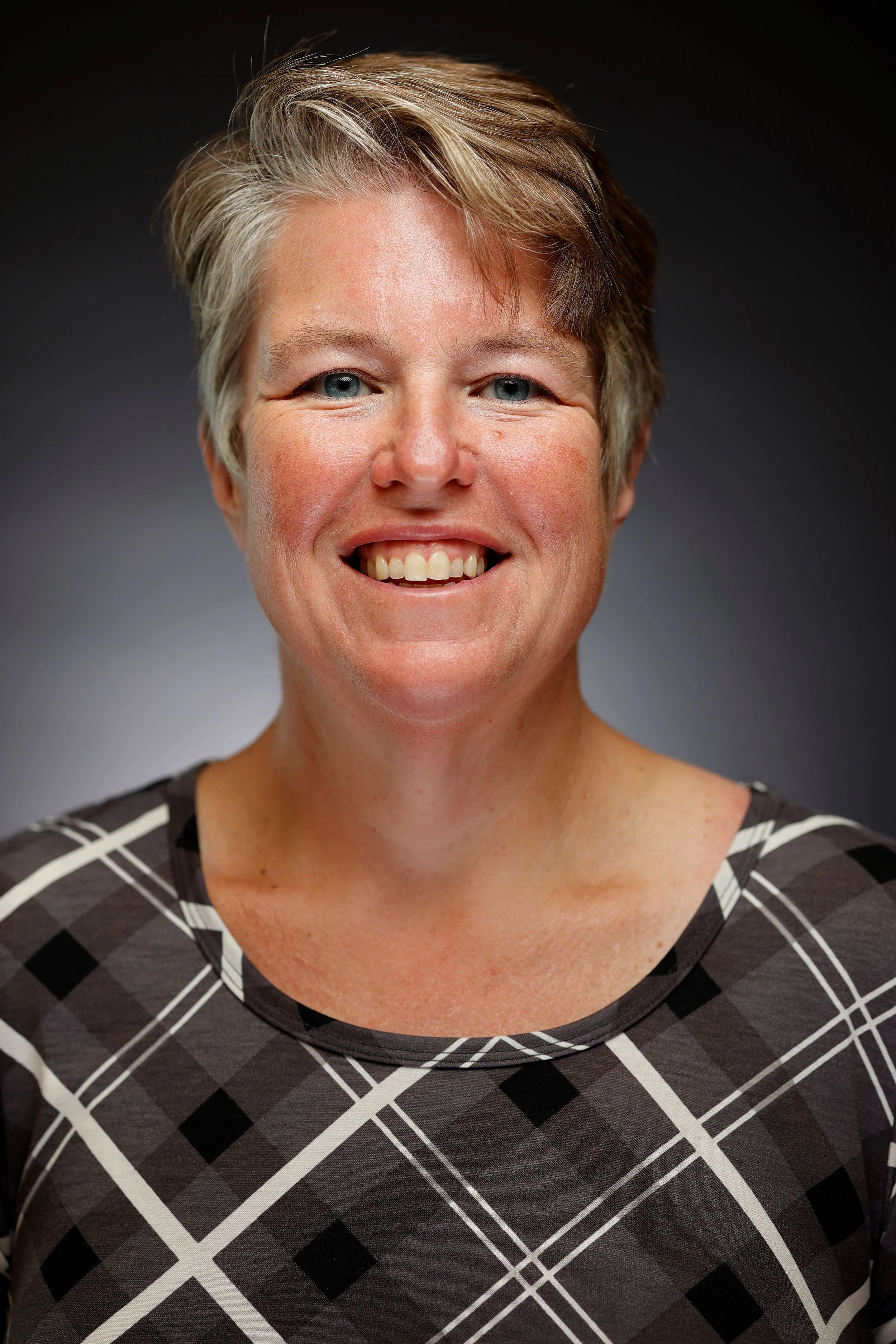 Kim Mackey smiling with short, light brown hair, blue eyes, wearing a black and white checkered top, against a dark gradient background.