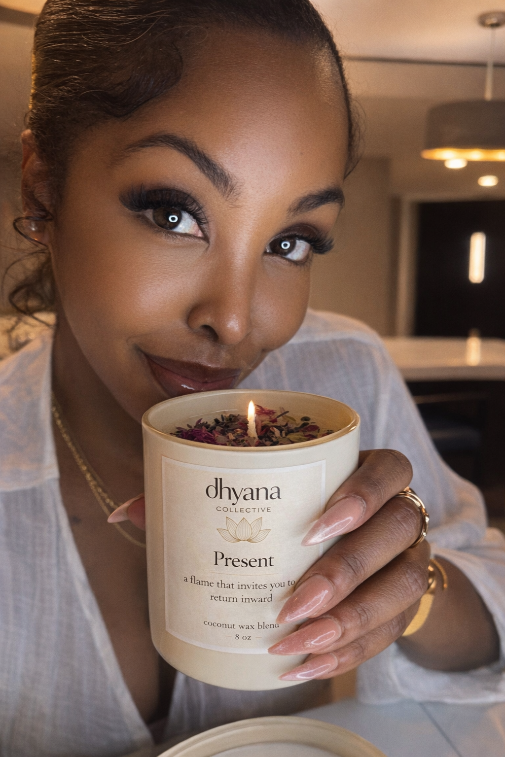A woman with styled hair and makeup holding a lit candle labeled "Present" from Dhyana Collective, with a warm and inviting indoor background.