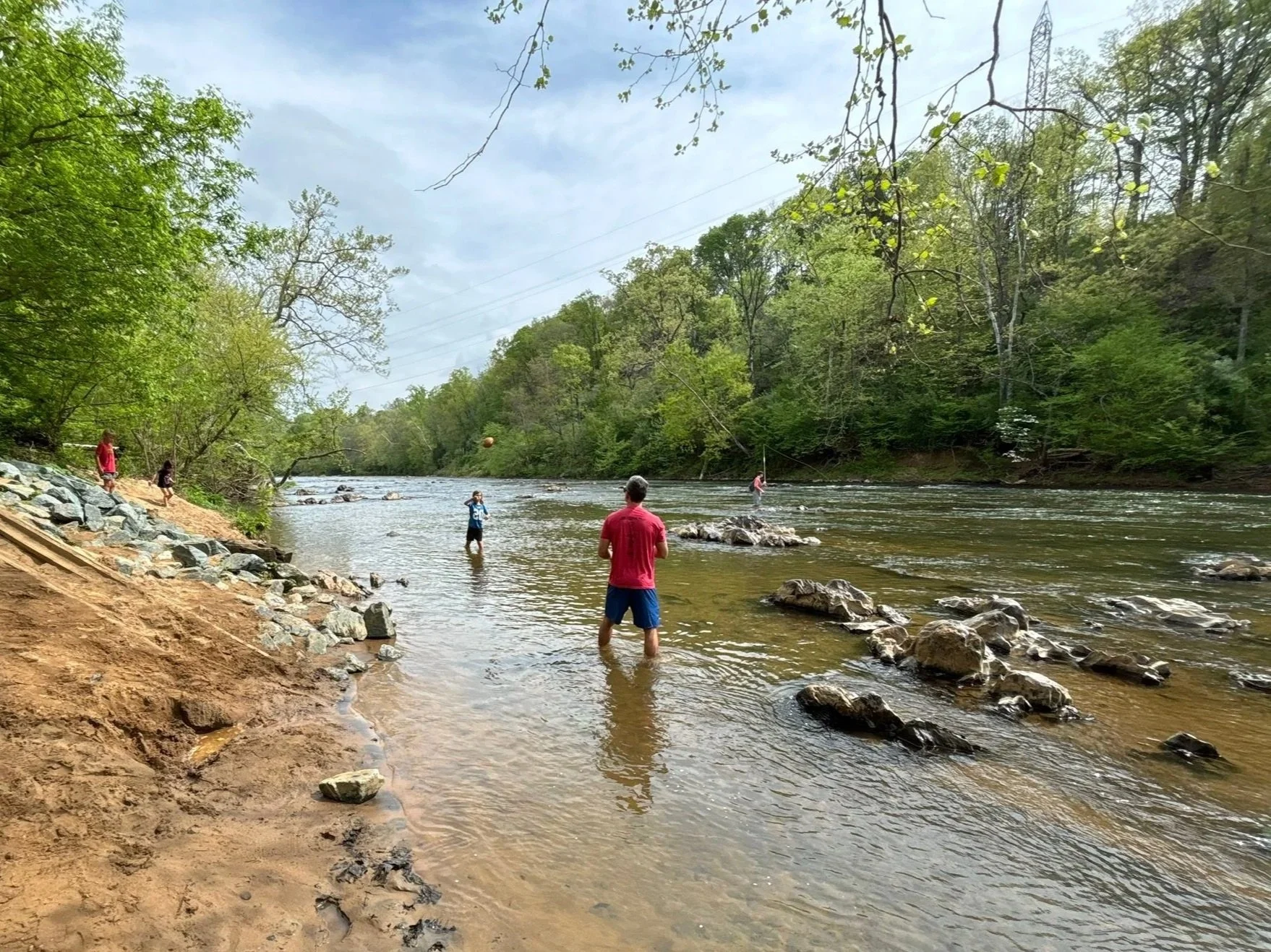 People wading and fishing in a river surrounded by green trees on a cloudy day.