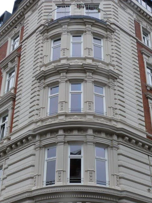 Mehrstöckiges Gebäude mit aufwendig gestalteten Fensterrahmen und dekorativen Ornamente im viktorianischen Stil, weiße Fassade, teilweise Ziegelmauerwerk visible.
