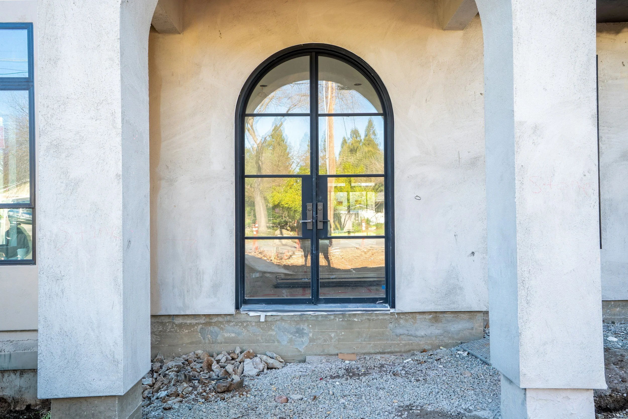 Fertiger Eingang mit schwarzen Metallrahmen-Tür und sandfarbenen Wänden, vor einer Baustelle mit Sand und Steinen.