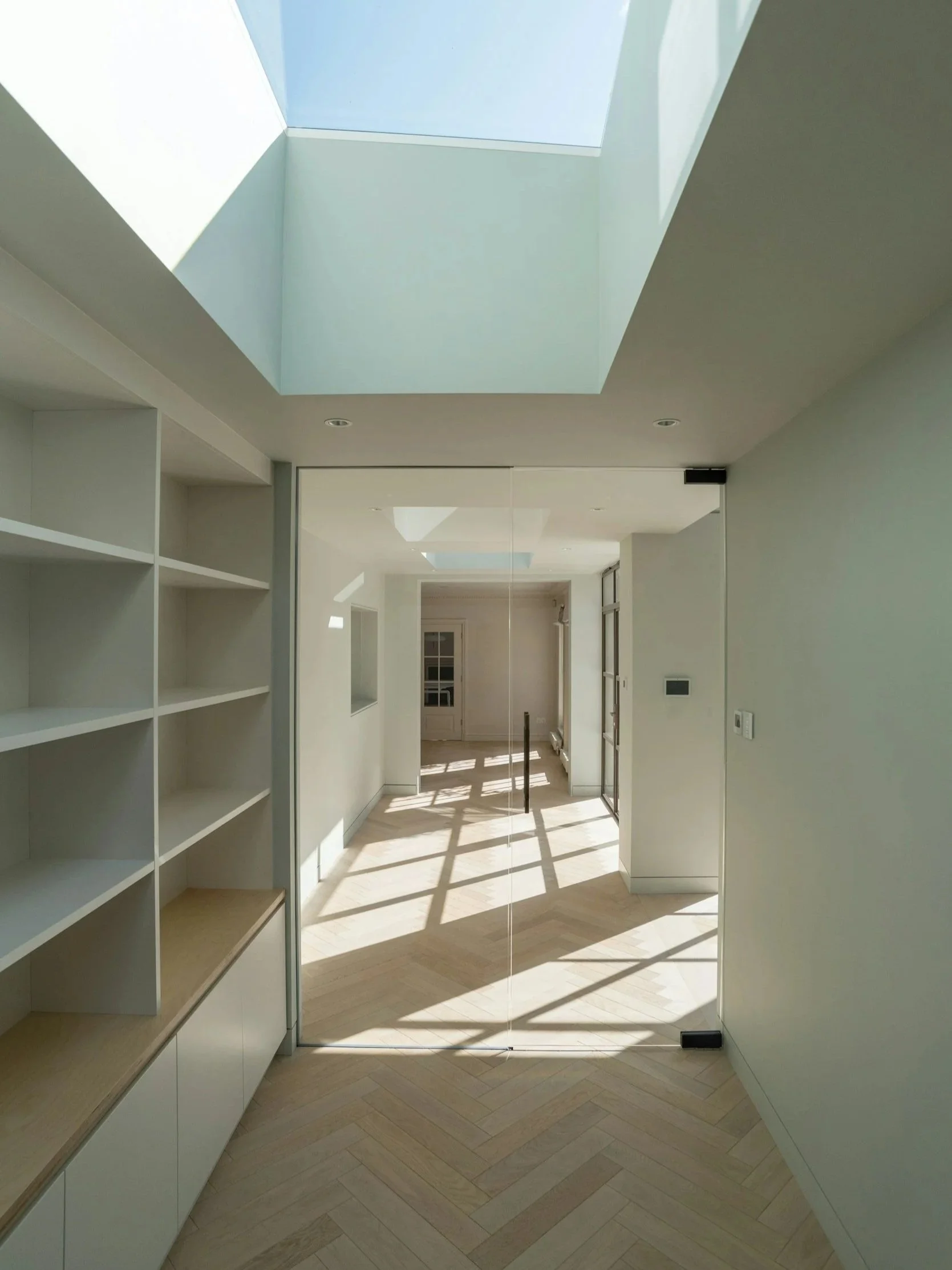 Ein heller, moderner Flur mit großem Dachfenster, das Sonnenlicht und Schatten auf den Boden wirft, durch eine Glastür sichtbar.