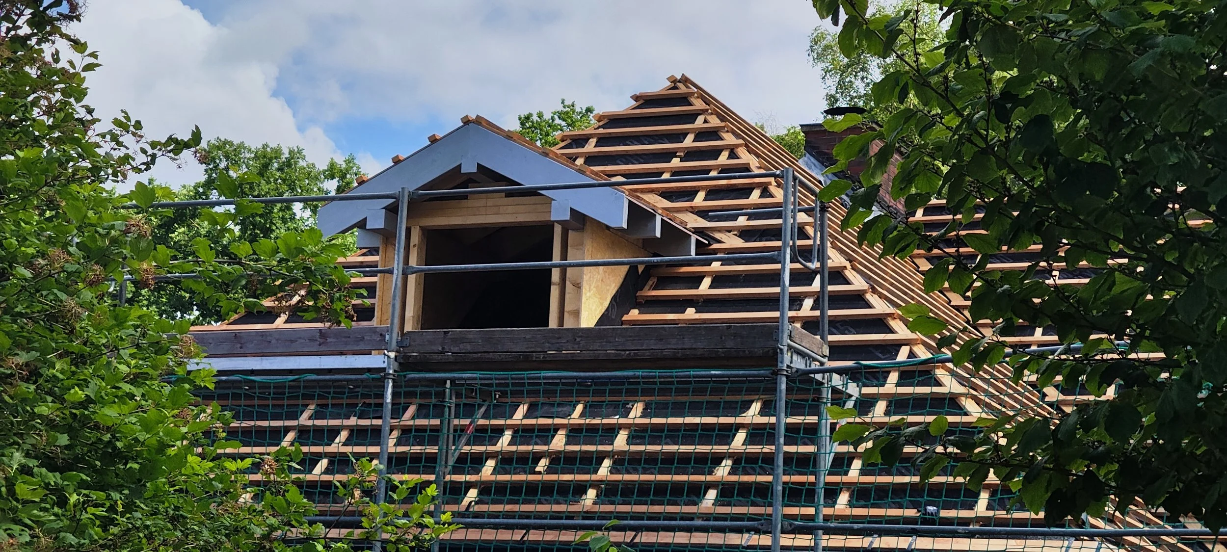 Zimmerdach im Bau mit Holzrahmen und Dachschindeln, umgeben von grünen Bäumen im Freien.