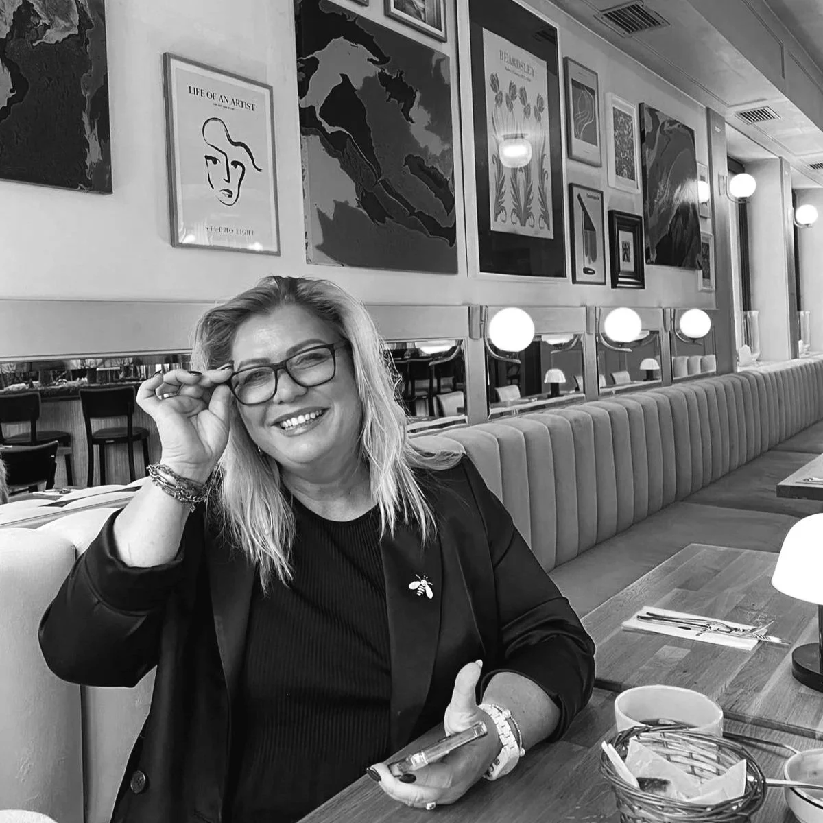 Frau lächelnd in einem Restaurant, trägt Brille und schwarzes Outfit, sitzt an einem Tisch mit Besteck und einer Tasse, Wand mit Kunstwerken im Hintergrund, Schwarz-Weiß-Foto.