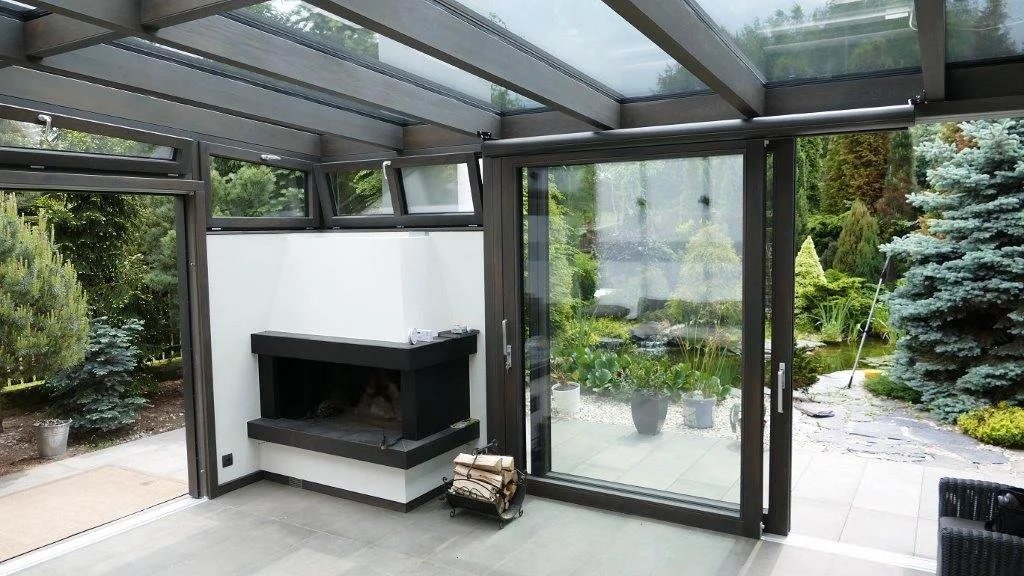Innenansicht eines Wintergartens mit Glastüren, einem Kamin und Blick auf einen gepflegten Garten mit Pflanzen und Bäumen.