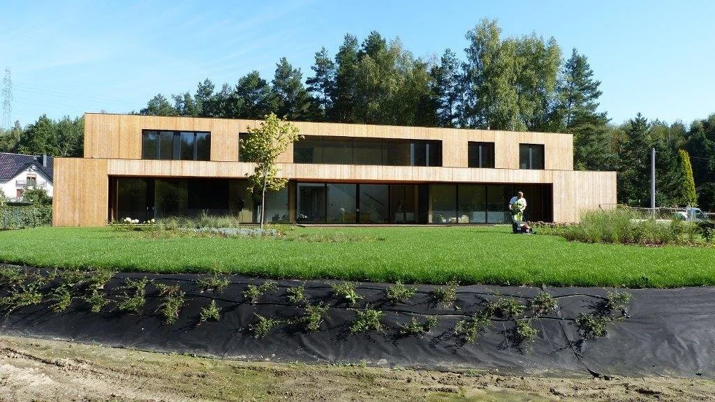 Modernes zweistöckiges Haus mit Holzfassade, große Fenster und grünem Garten im Vordergrund, bewaldete Hügel im Hintergrund.