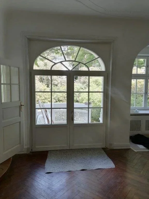 Helles Zimmer mit großen Glastüren zum Balkon, Holzfußboden, Fenster auf der rechten Seite, kleiner Teppich vor den Türen.