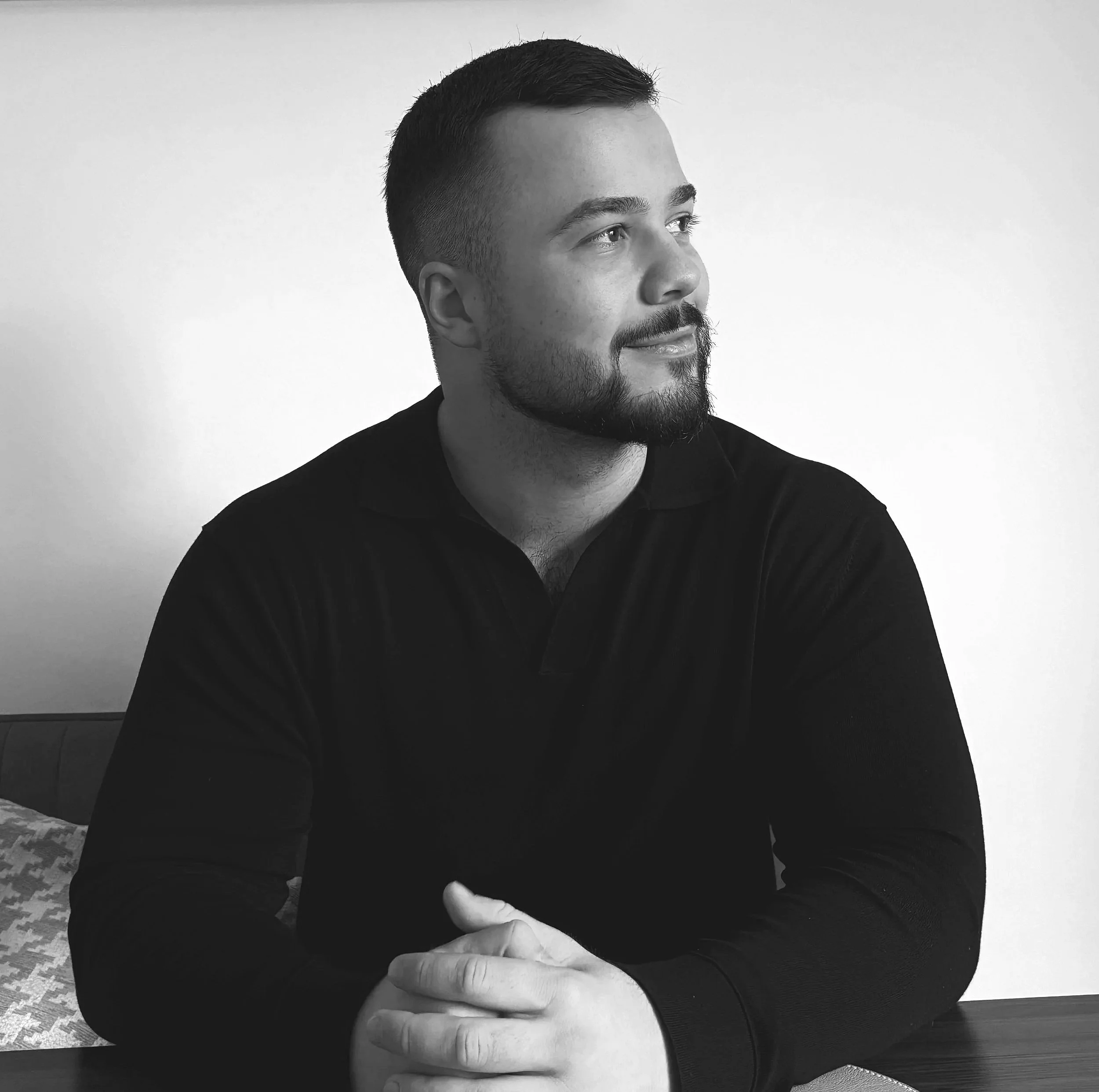 Schwarz-weiß-Fotografie eines jungen Mannes mit Bart, der nach rechts schaut, an einem Tisch sitzt und die Hände hält.