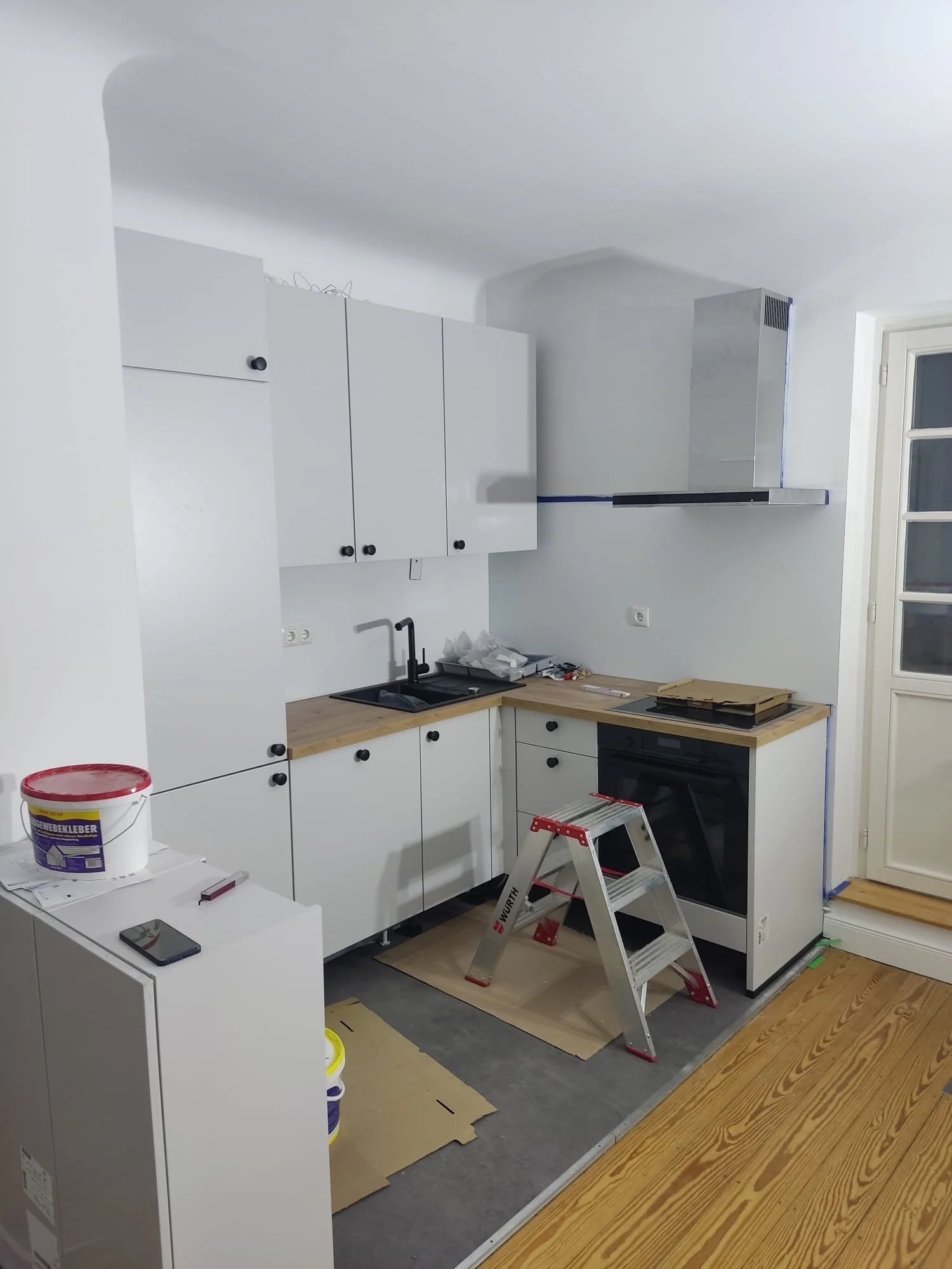 Küche in Renovierung, mit weißen Schränken, einem schwarzen Ofen, einem schwarzen Wasserhahn, einer Dunstabzugshaube, einem roten Stehleiter, Bauarbeitenutensilien und Verpackungen auf den Oberflächen.
