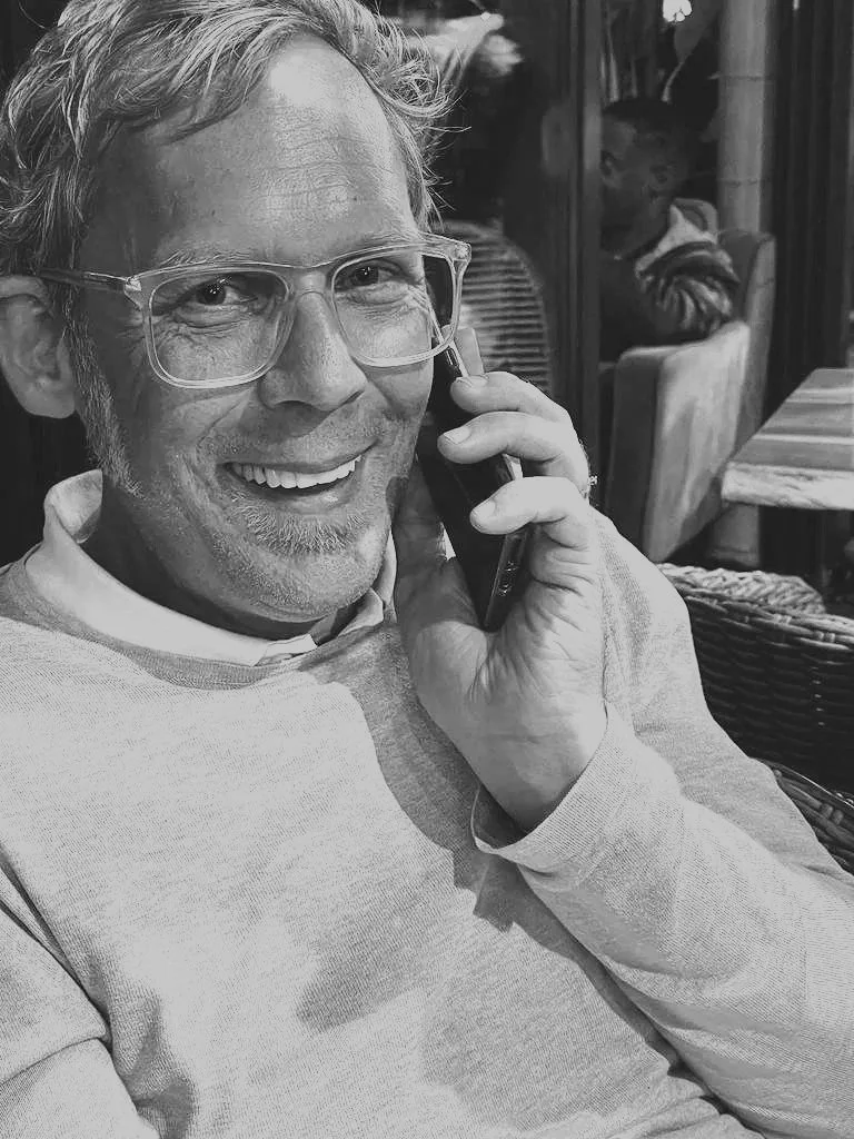 Ein Mann mit Brille und grauem Bart lächelt, während er telefoniert, im Hintergrund weitere Personen im Café.