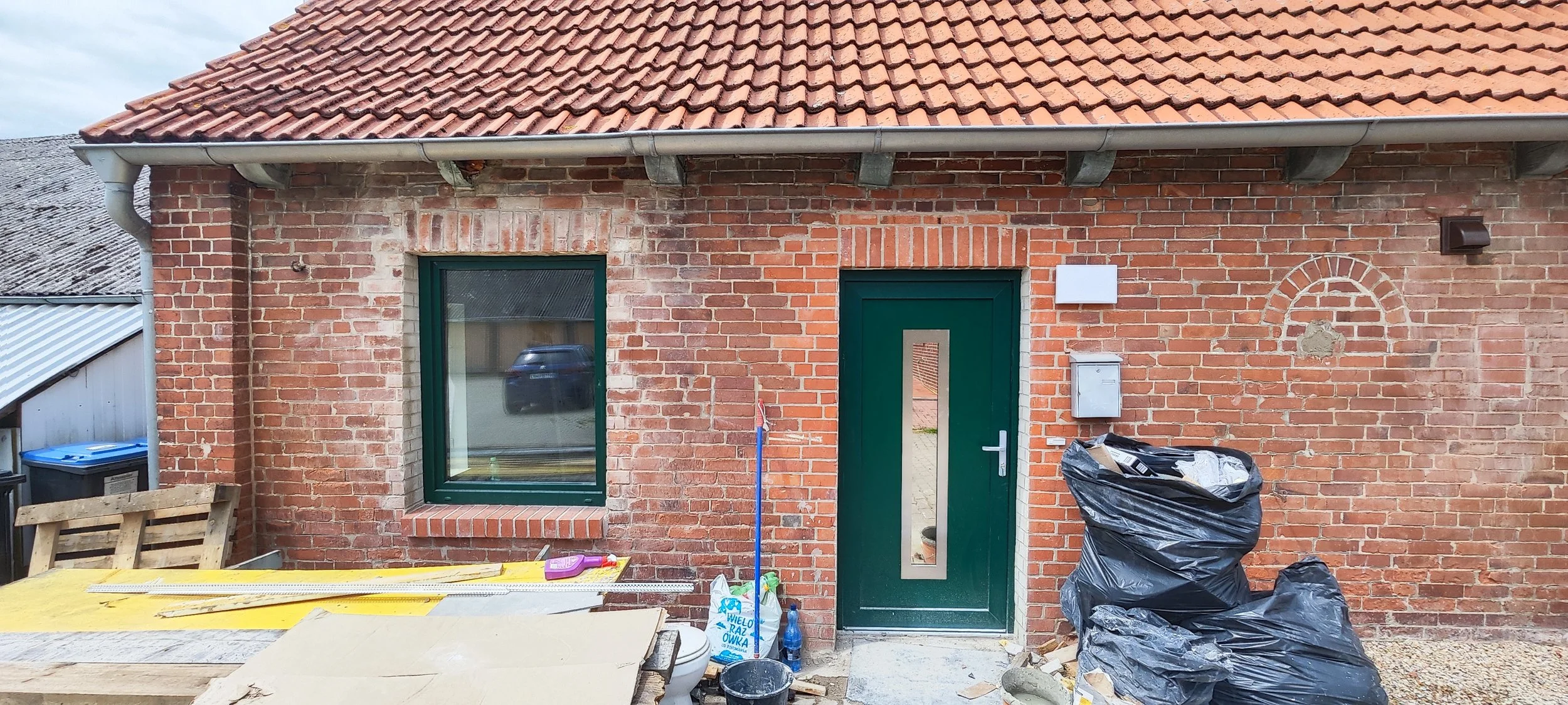 Eine rotrebrannte Hauswand mit grünem Fenster und Tür. Im Vordergrund befinden sich Baustellenmaterialien und Müllsäcke.