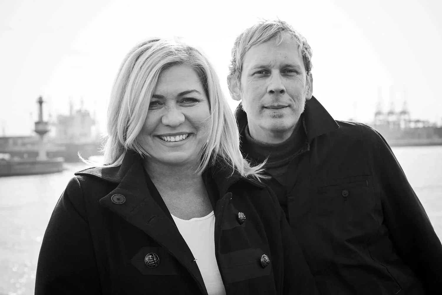 Ein schwarz-weiß Foto eines lachenden Frauen und eines Mannes, die am Wasser stehen, mit einem Hafen im Hintergrund.