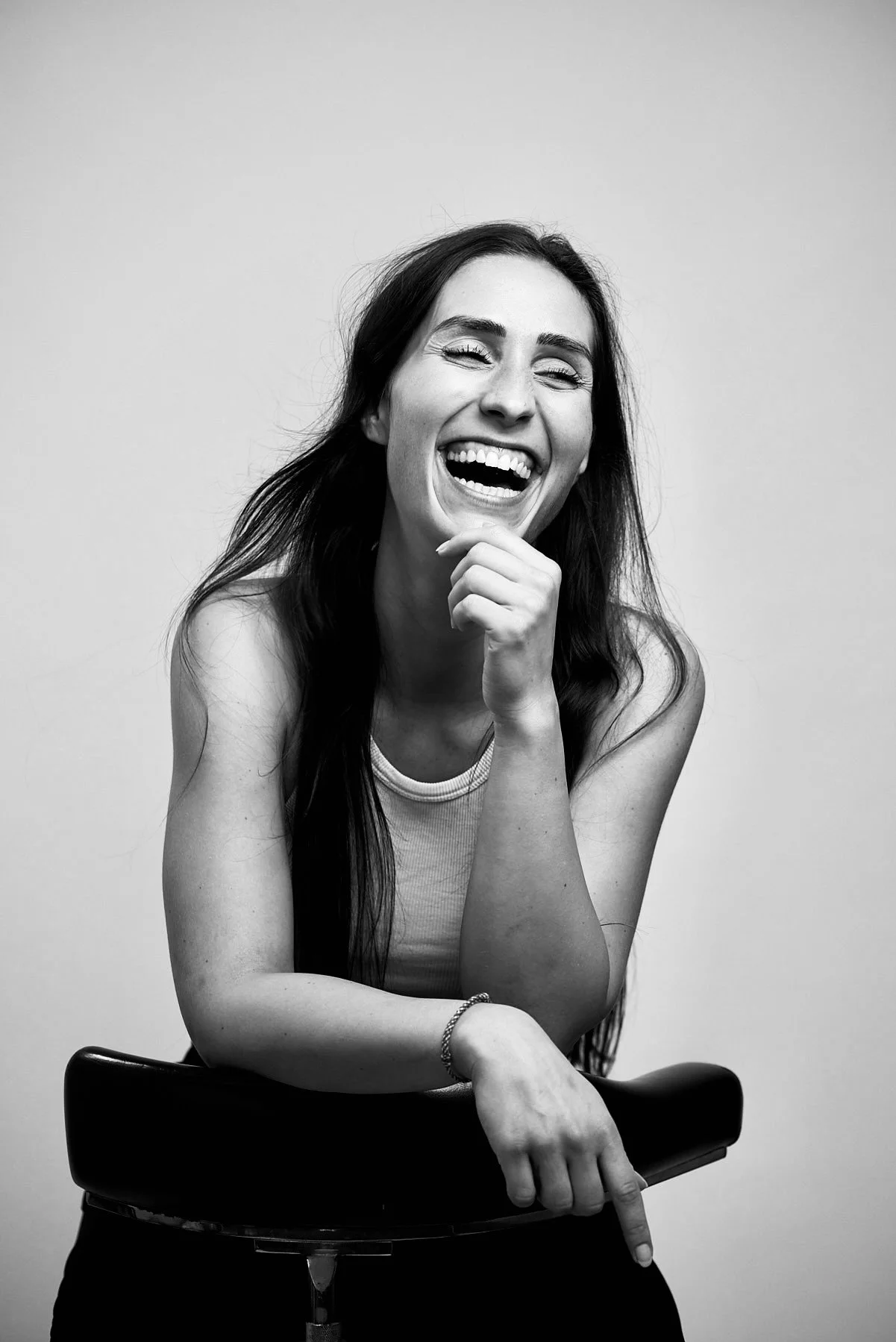 Schwarzweiß-Foto einer lachenden Frau mit langen dunklen Haaren, die auf einem Stuhl sitzt, gegen einen einfachen hellen Hintergrund.