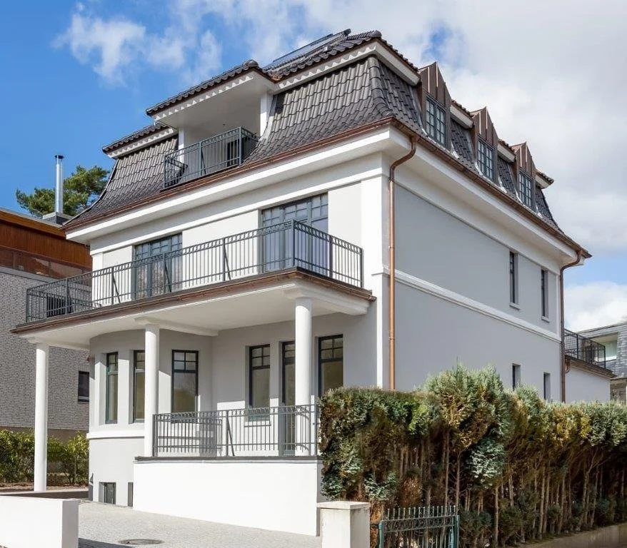 Mehrstöckiges weißes Haus mit Balkon und dunklem Schieferdach, umgeben von Bäumen und einem Zaun.