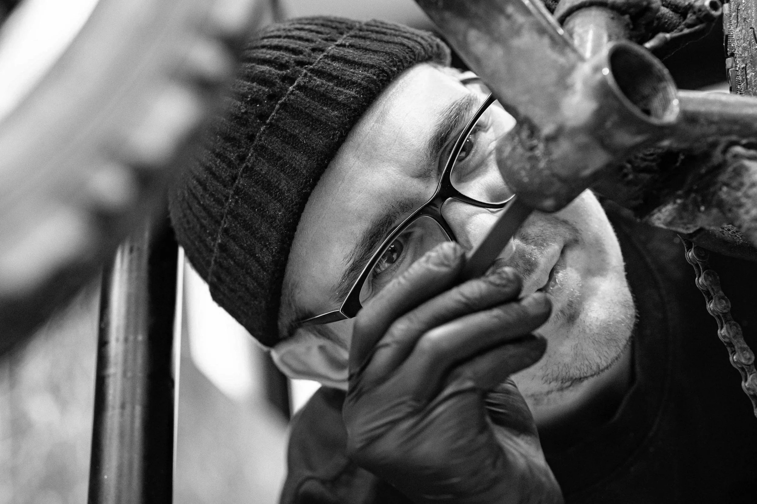 Ein junger Mann trägt eine dunkle Mütze und Brille, arbeitet an einem Fahrrad, und nutzt ein Werkzeug. Er ist in einer Werkstatt oder an einem Reparaturort für Fahrräder.