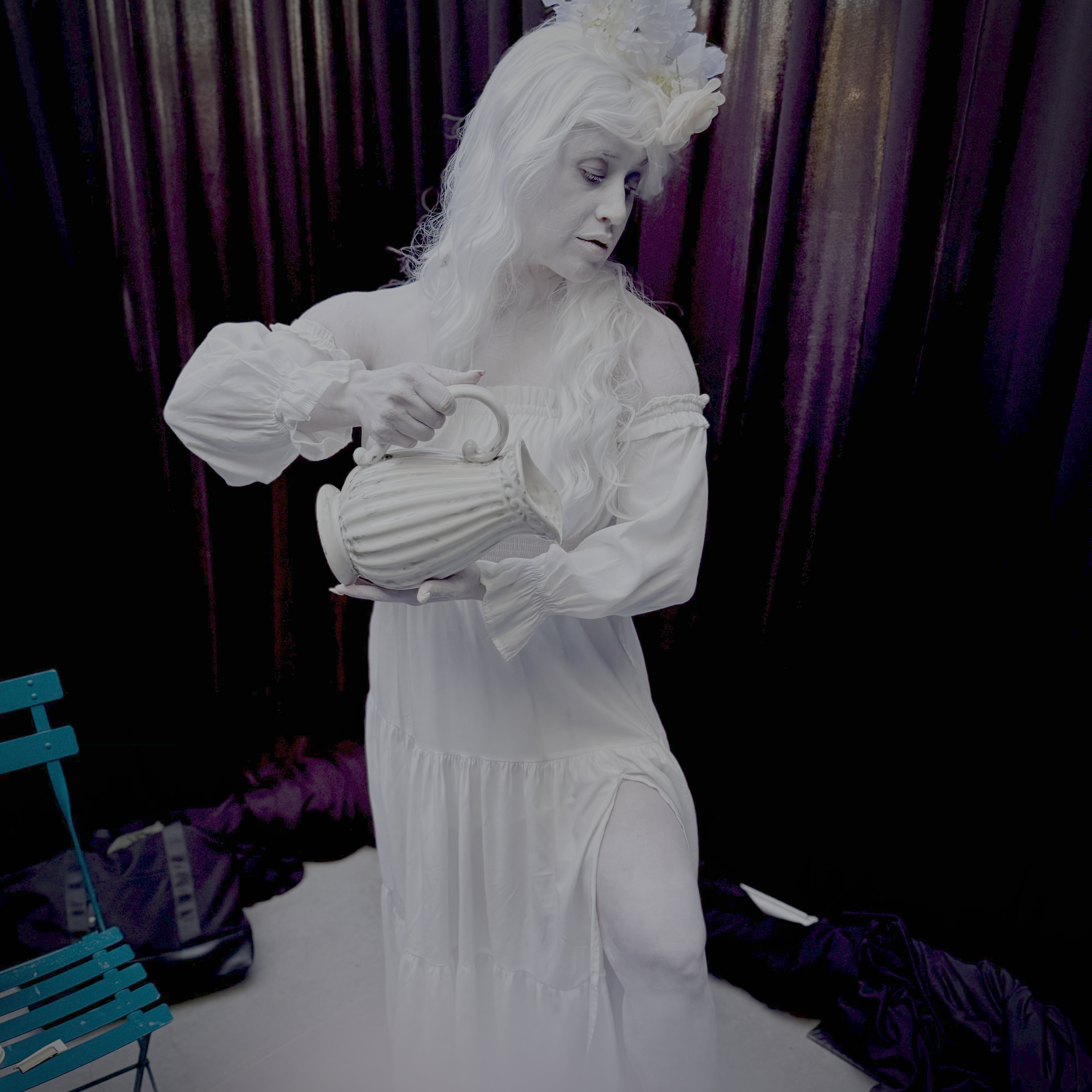 A woman dressed as a statue with white painted face and curly white hair, wearing a white dress and a floral headpiece, pouring water from a white ceramic pitcher.