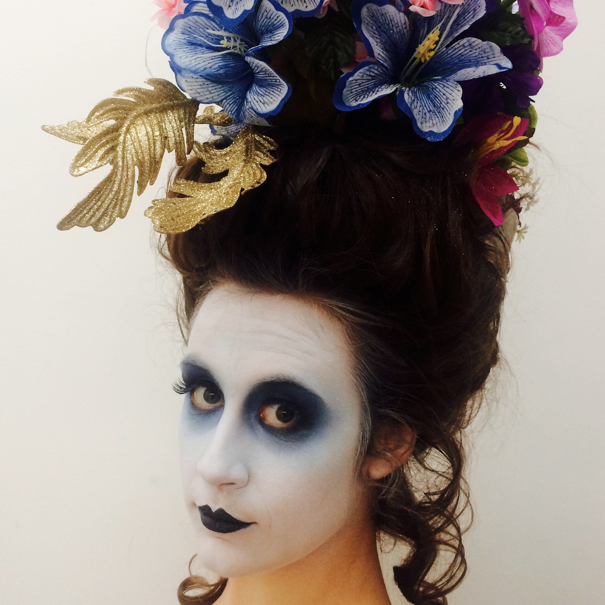 A woman with skull-like makeup, blue eyeshadow, and black lipstick, wearing a tall headpiece with blue, pink, and purple flowers, and gold decorative leaves.