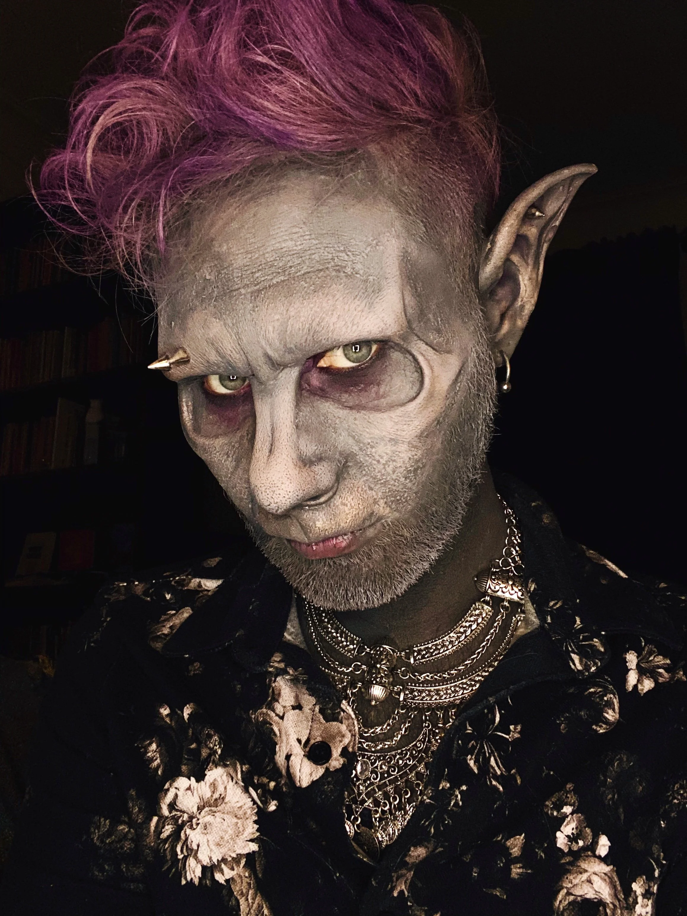 Person with elf-like ears, silver face paint, purple hair, multiple layered necklaces, and wearing a floral patterned shirt.