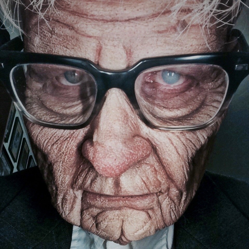 A close-up of an elderly man wearing large black glasses, with deeply wrinkled skin, gray hair, and a serious expression.