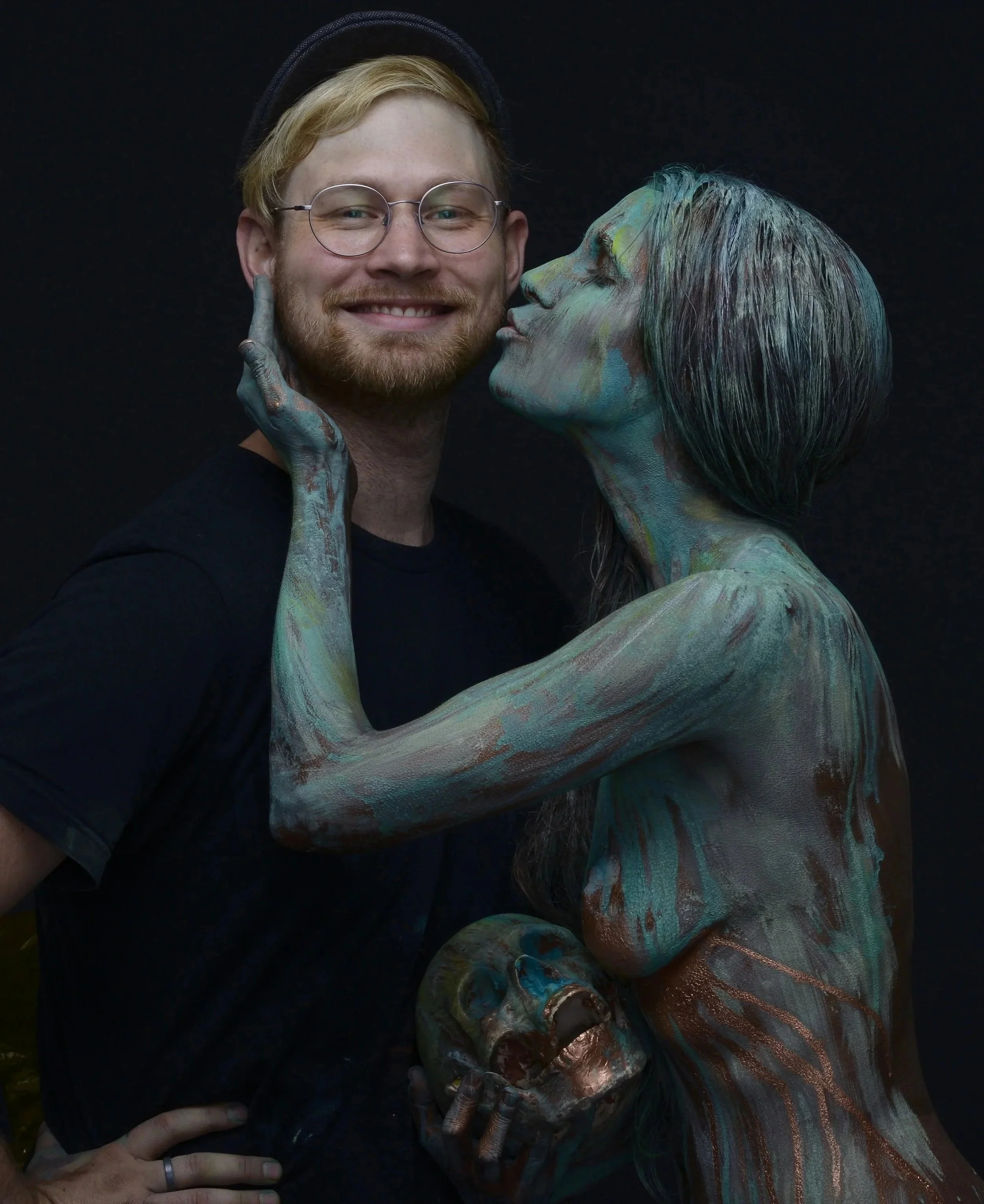 A man with glasses and a hat smiling as a woman with painted body and face is kissing his cheek, holding his chin with her hand. The woman has paint in shades of green, blue, and bronze covering her body and face, and she holds a painted skull in her hand.
