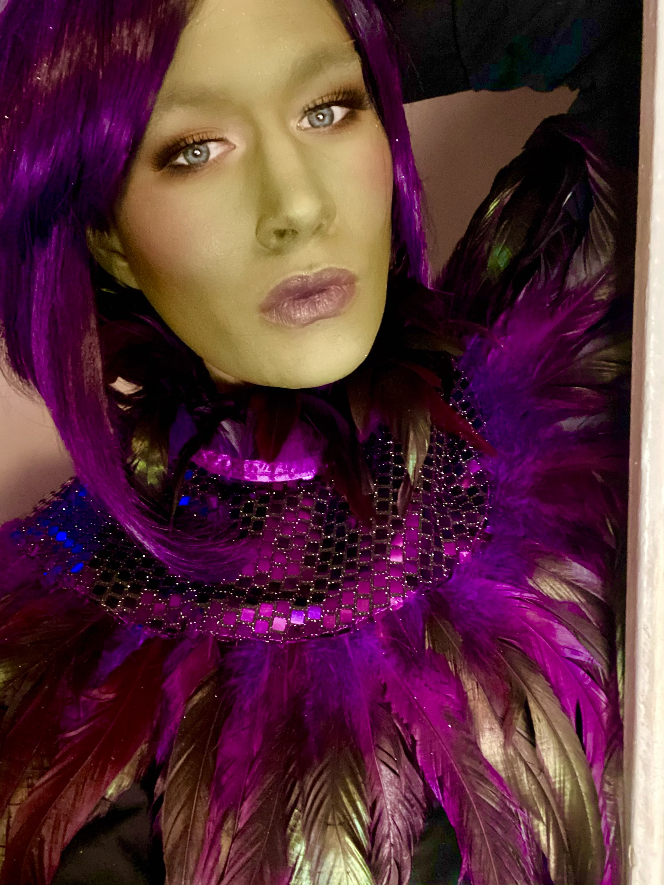 A woman with purple hair and bold makeup wearing an elaborate costume with purple feathers and black mesh adorned with small reflective squares.