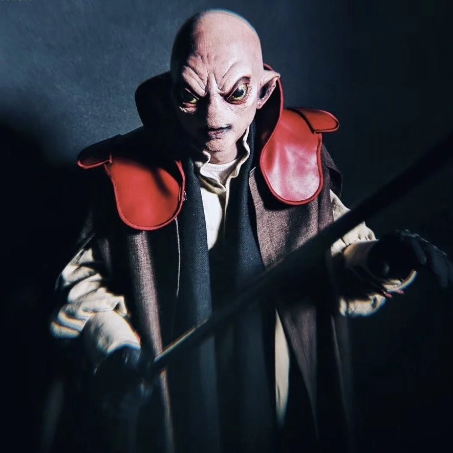 A humanoid alien with large eyes and bald head dressed in a brown and black robe with red shoulder pads, holding a staff, in a dark setting.