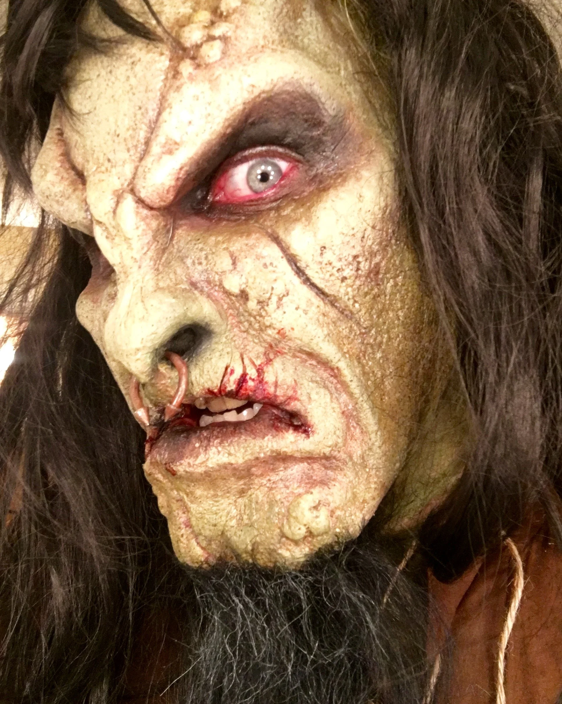 Close-up of a person with zombie makeup, including greenish rugged skin, a scar on the forehead, dark eye makeup, a blood-red eye, and wearing a nose ring, with long black hair and a beard.