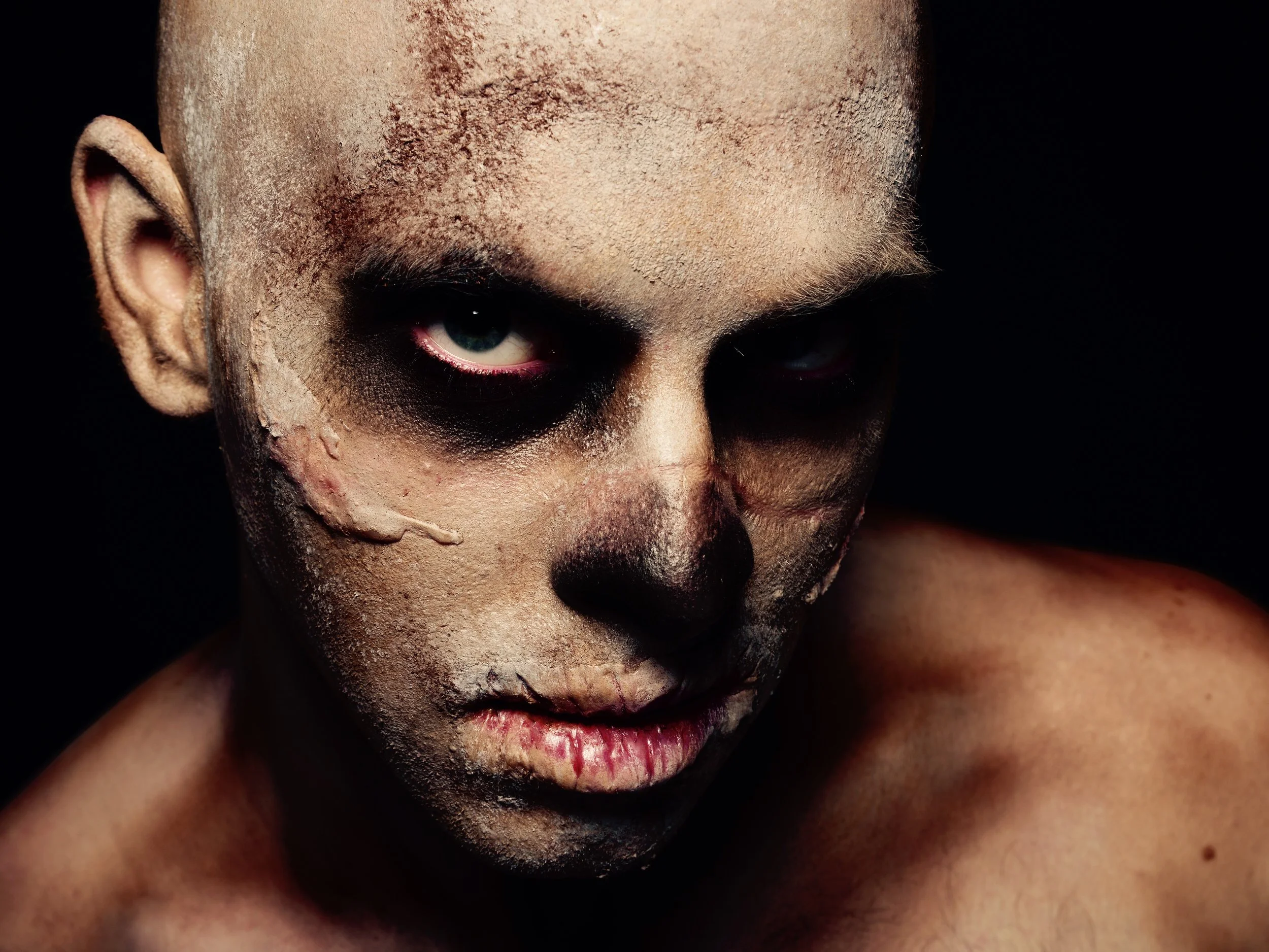 Close-up of a person with zombieskin makeup, dirty and cracked face paint, and intense eyes, against a dark background.
