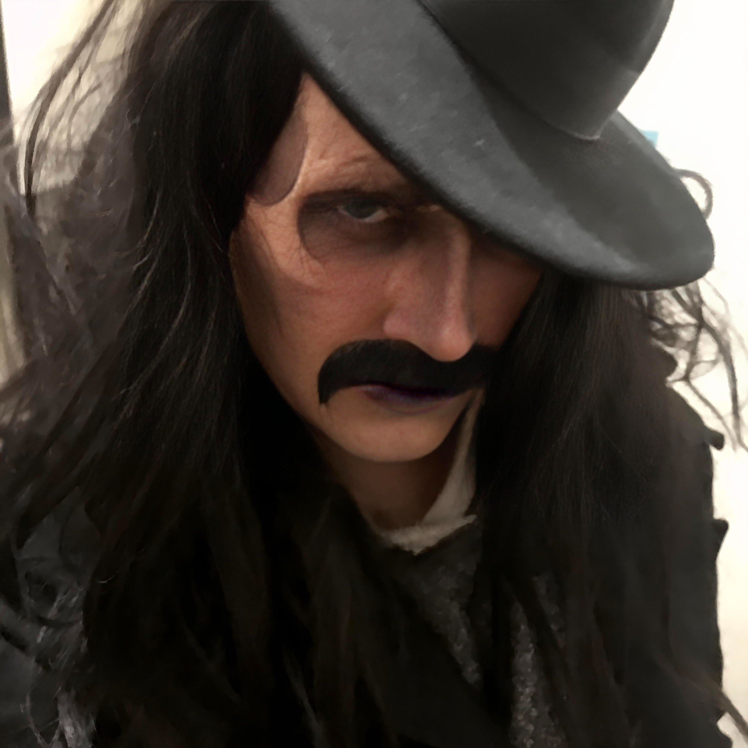 Person dressed as a classic villain or magician, wearing a black hat and coat, with a painted mustache, long dark hair, and intense expression