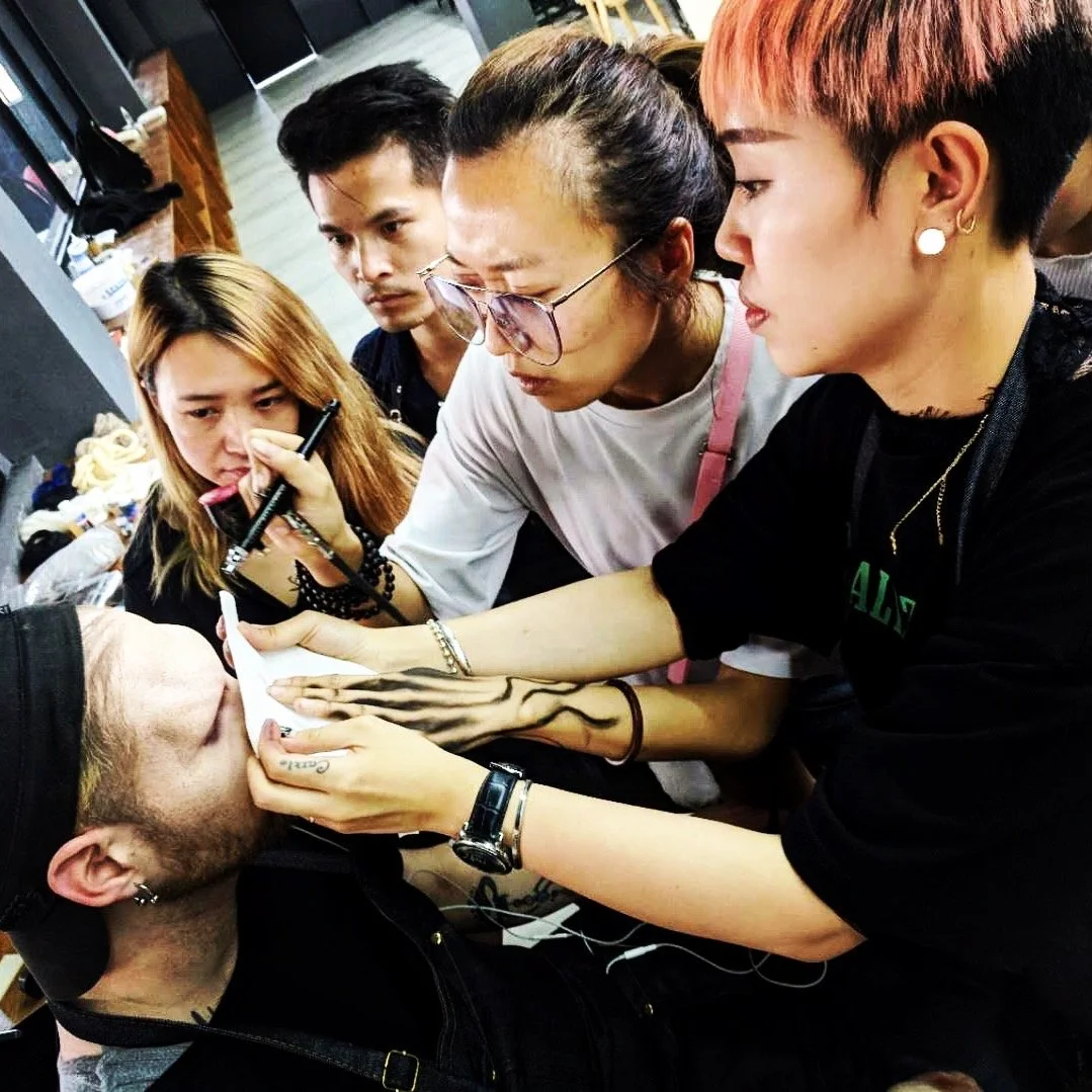 Group of five young adults gathered around, with one person applying makeup or face paint on another person, while others observe closely.
