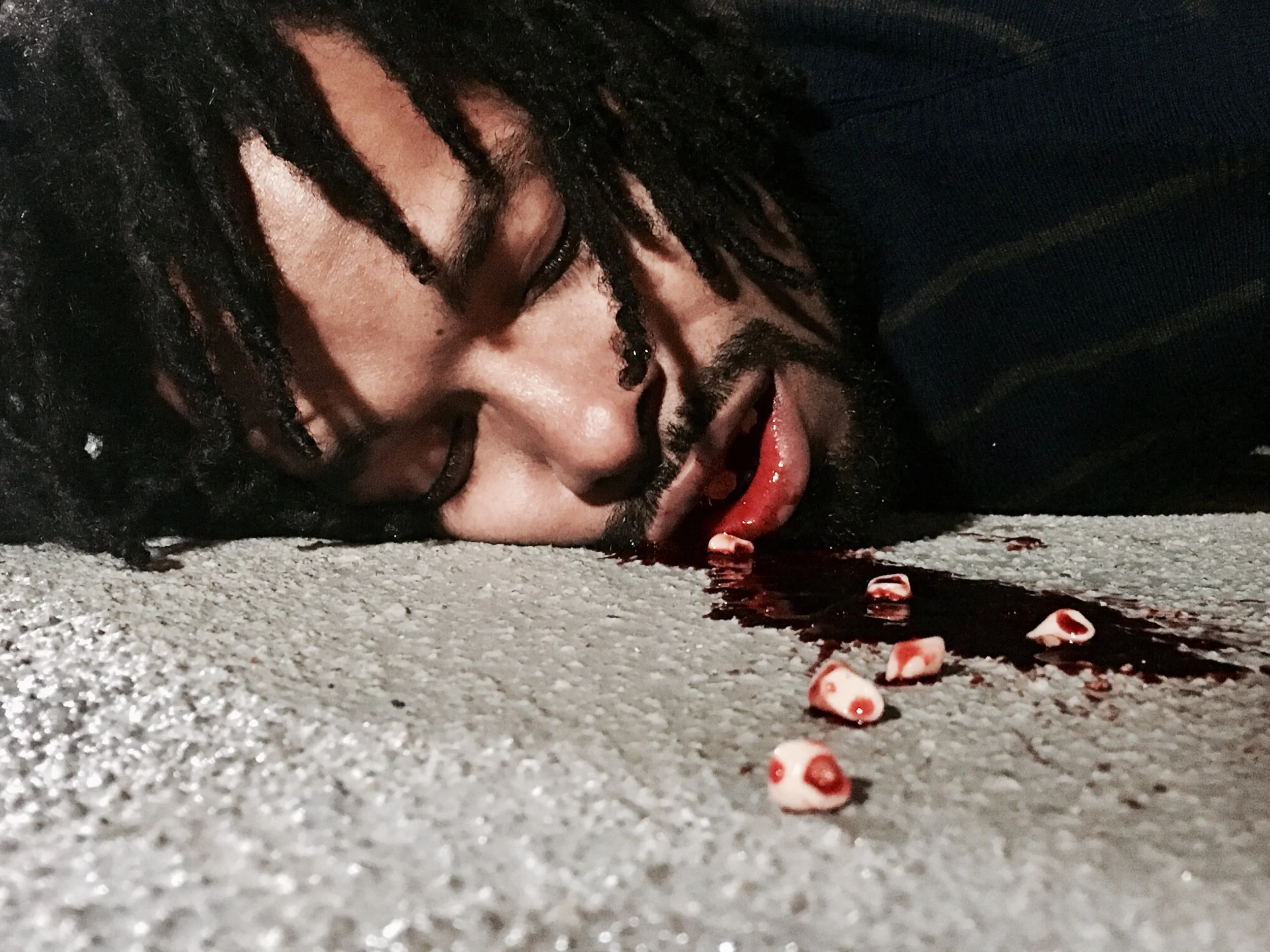 Close-up of a man lying on the ground with blood and small red and white pills spilled near his face.