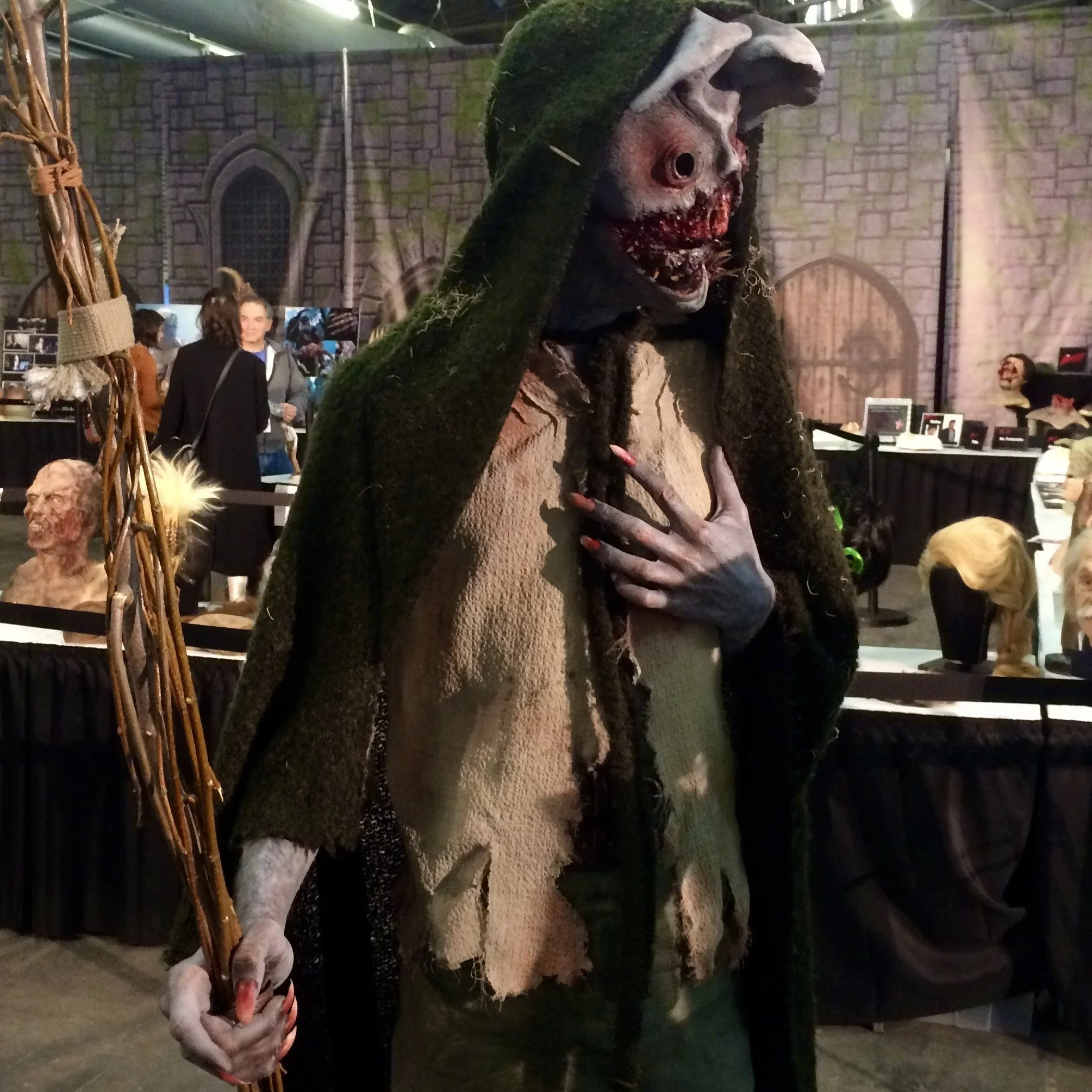 A person dressed as a creature with a zombie-like face, tattered clothing, and dreadlocks, holding a stick with twigs, at a costume or Halloween event.