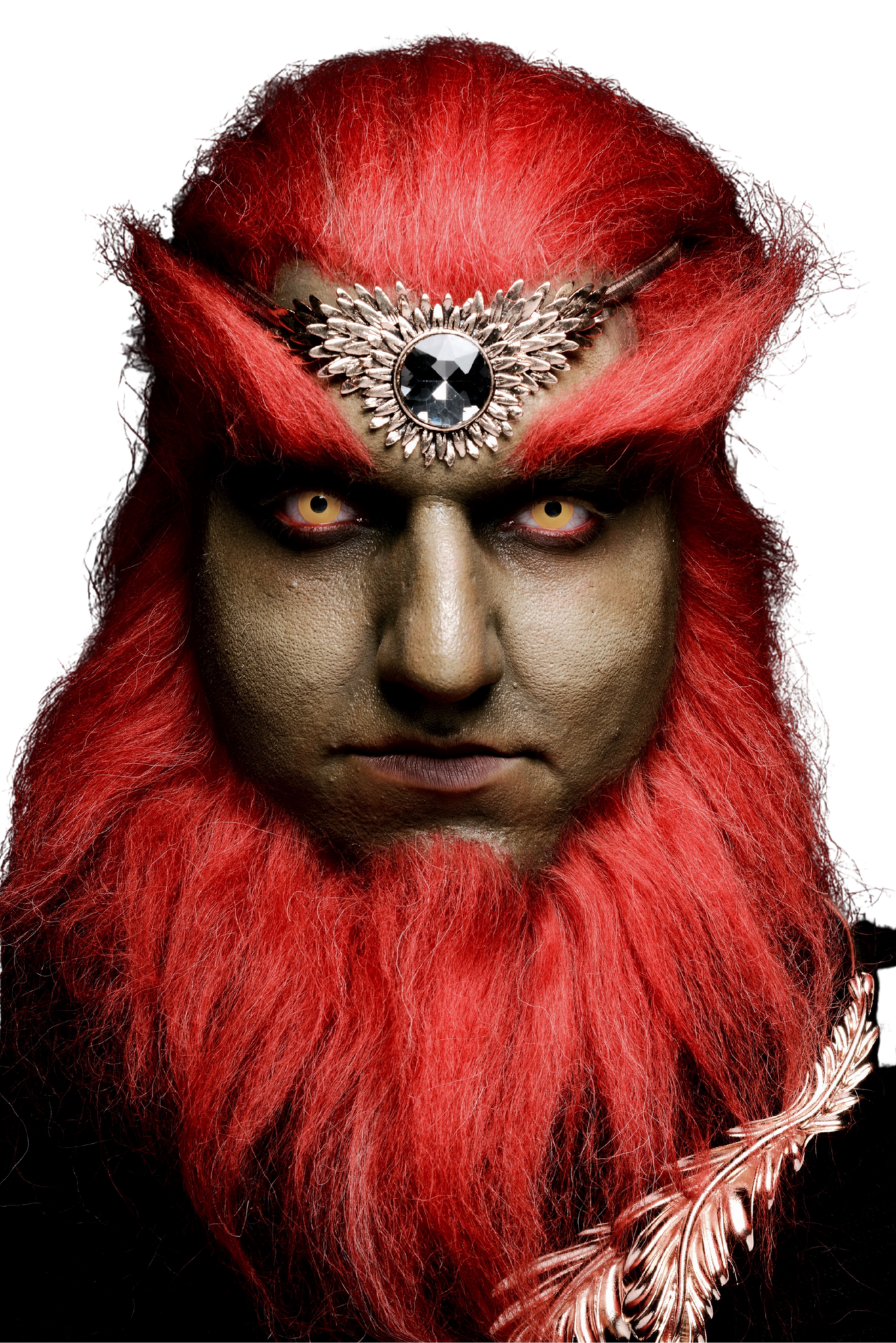 Person with dark makeup, red hair and beard, wearing an ornate silver headpiece with a large black gemstone, against a black background.