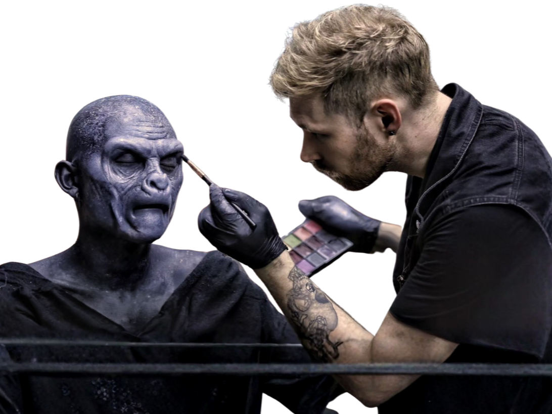 Makeup artist applying special effects makeup to a person's face, creating a horror-like character with dark, cracked skin and hollow eyes.