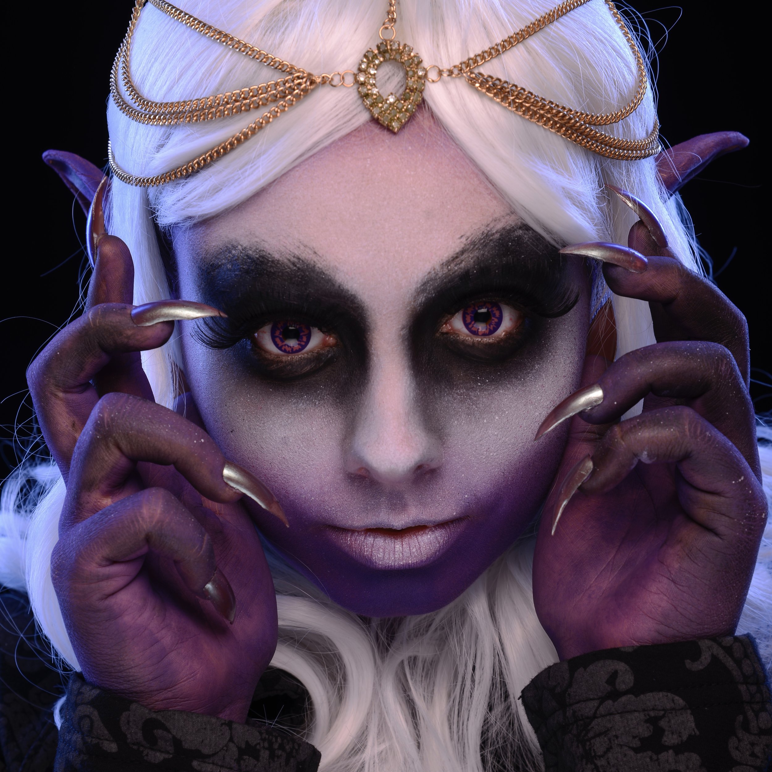 A person with fantasy makeup, white wig, gold jewelry, purple hands, and contacts with a swirling pattern, creating a supernatural look.