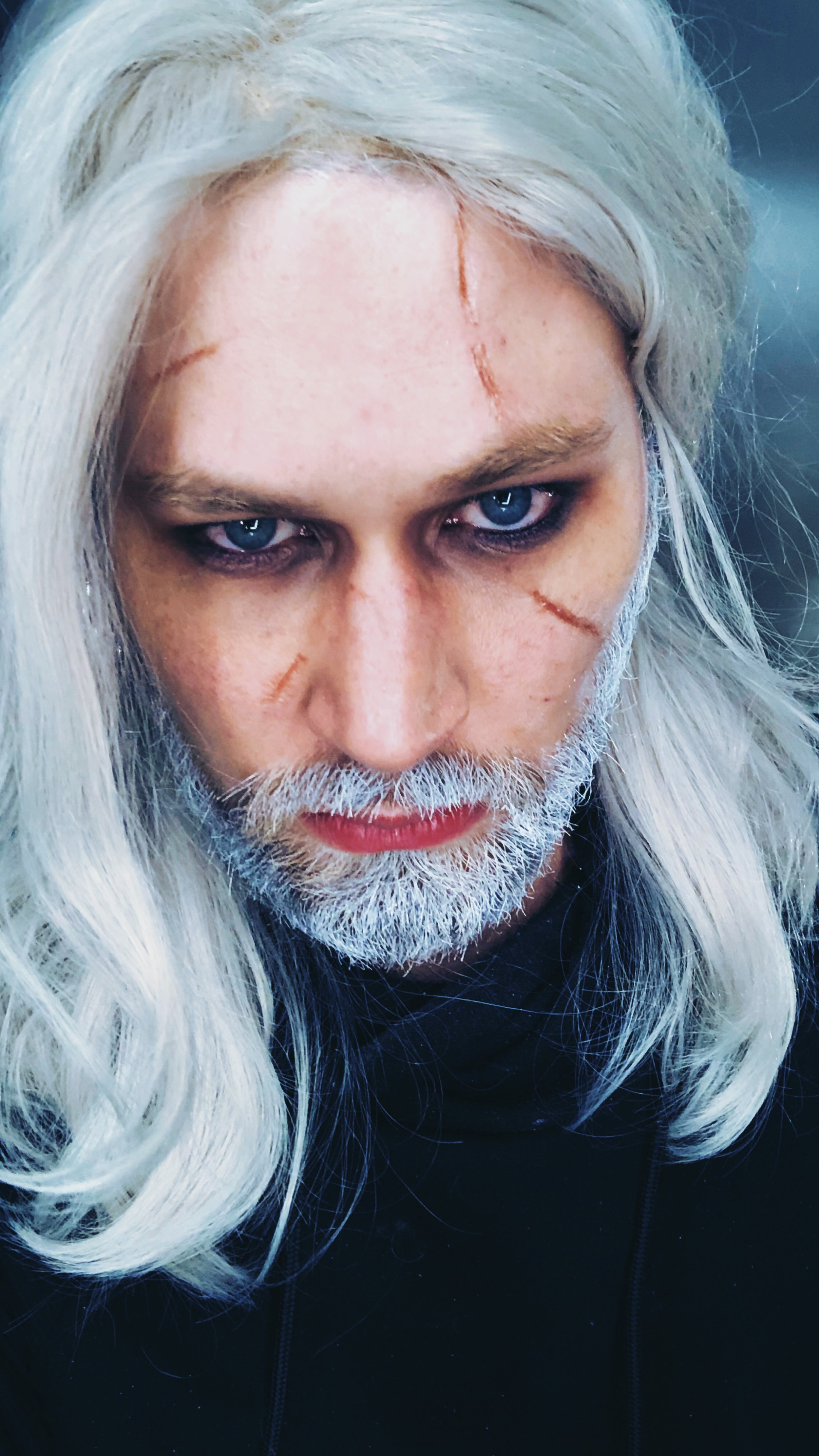 Close-up of a man with long, white hair, blue eyes, and a beard, looking intensely at the camera with scars on his face.