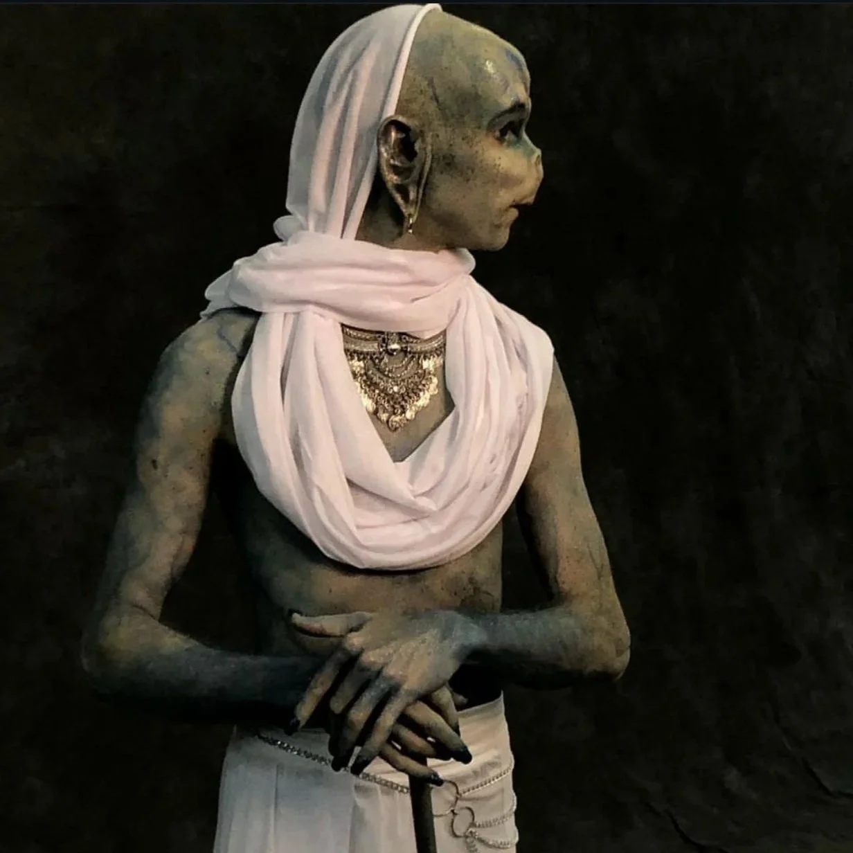 Statue of a woman with traditional jewelry, wearing a white headscarf and draped fabric, set against a dark background.