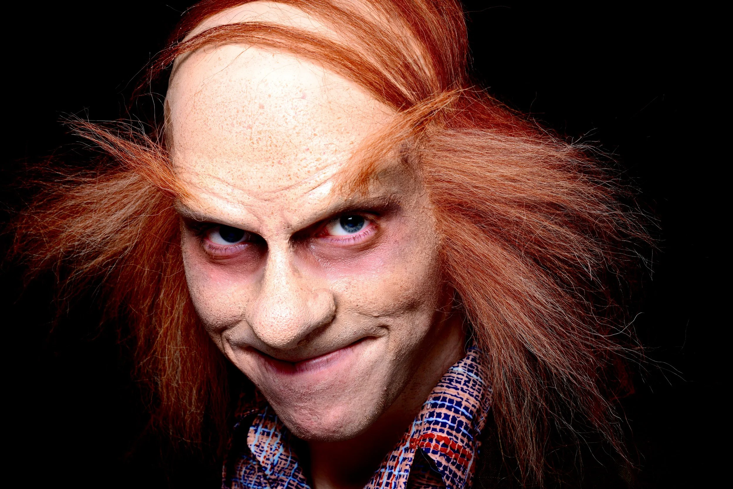Close-up of a man with wild, red hair, wearing a checkered shirt, with a mischievous expression against a black background.
