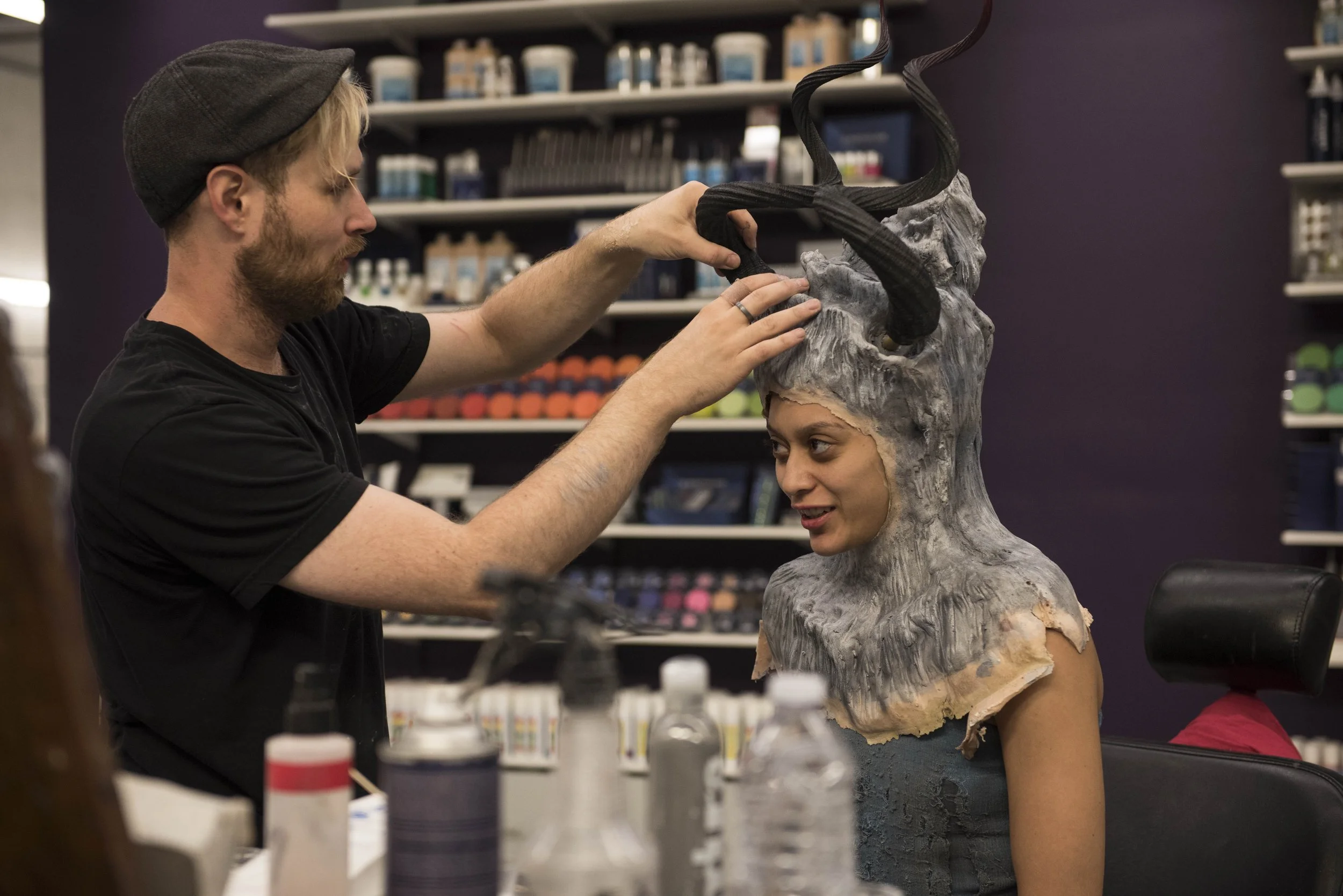 A man is putting a large, elaborate headpiece resembling a fantasy or creature character on a woman’s head. The headpiece covers her head and shoulders and has dark, tentacle-like extensions. The woman is smiling and looking at the man, and they are in a room with shelves of supplies in the background.