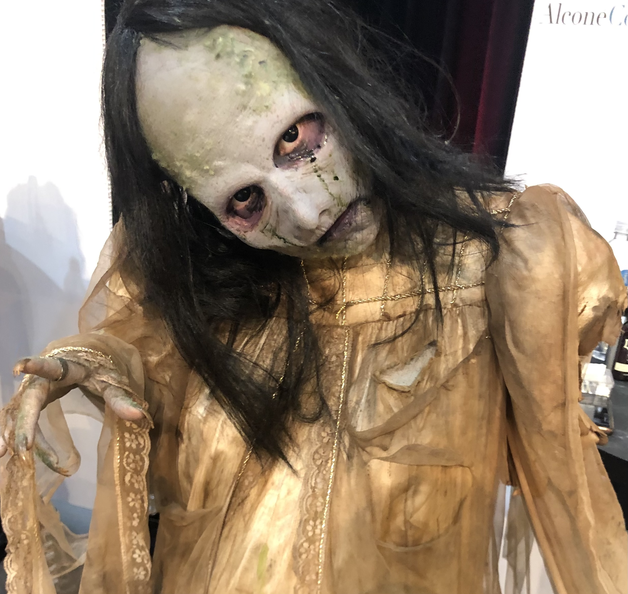 Person in a horror costume wearing a weathered, creepy mask with black hair, dressed in a tattered beige dress, and reaching out with a bony hand.